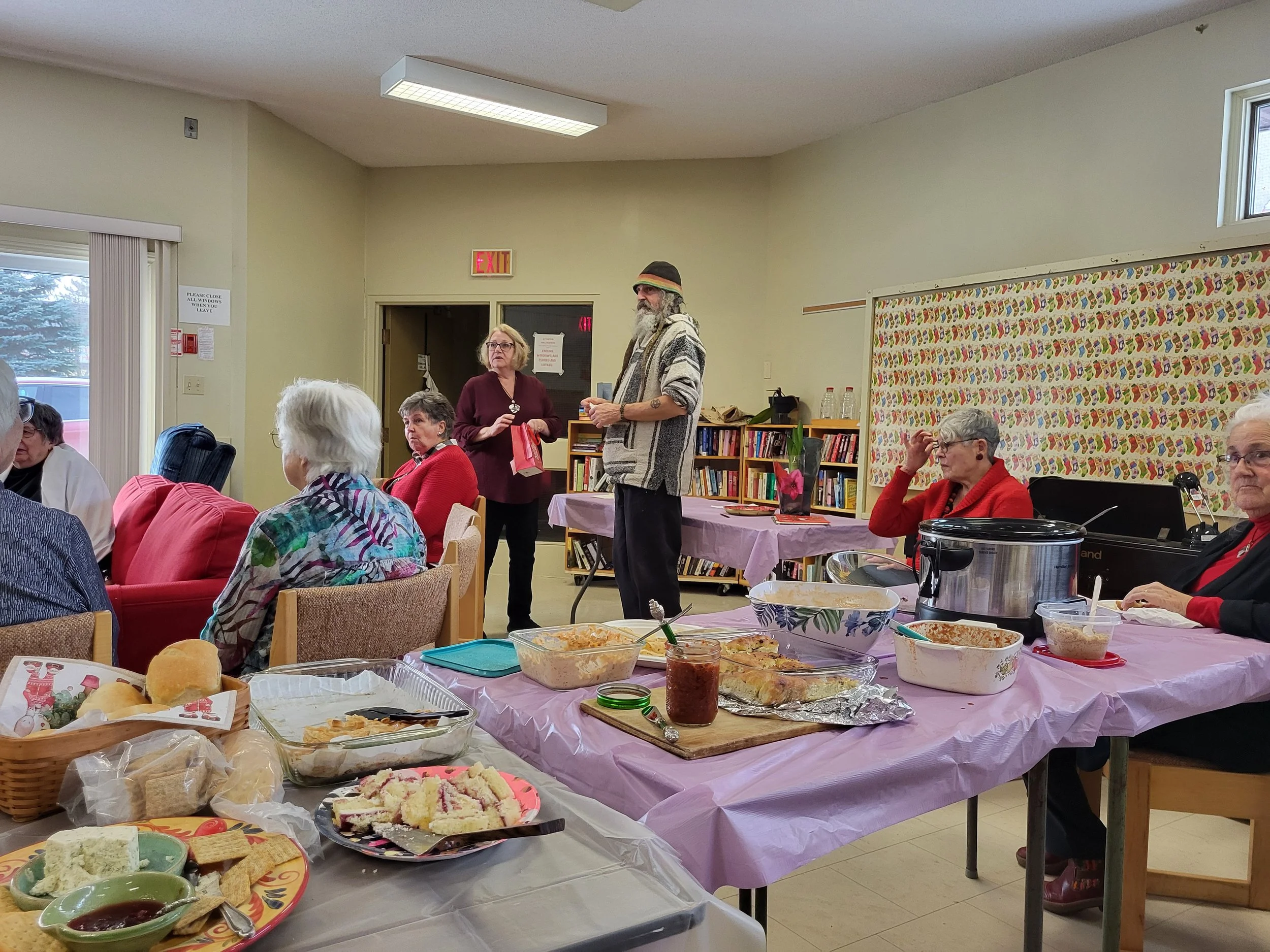 A group of people gathered in a room for a social event, with a table of food including sandwiches, cake, and snacks in the foreground. Several women, mostly elderly, are seated while a man stands and speaks, with additional women sitting and listening.