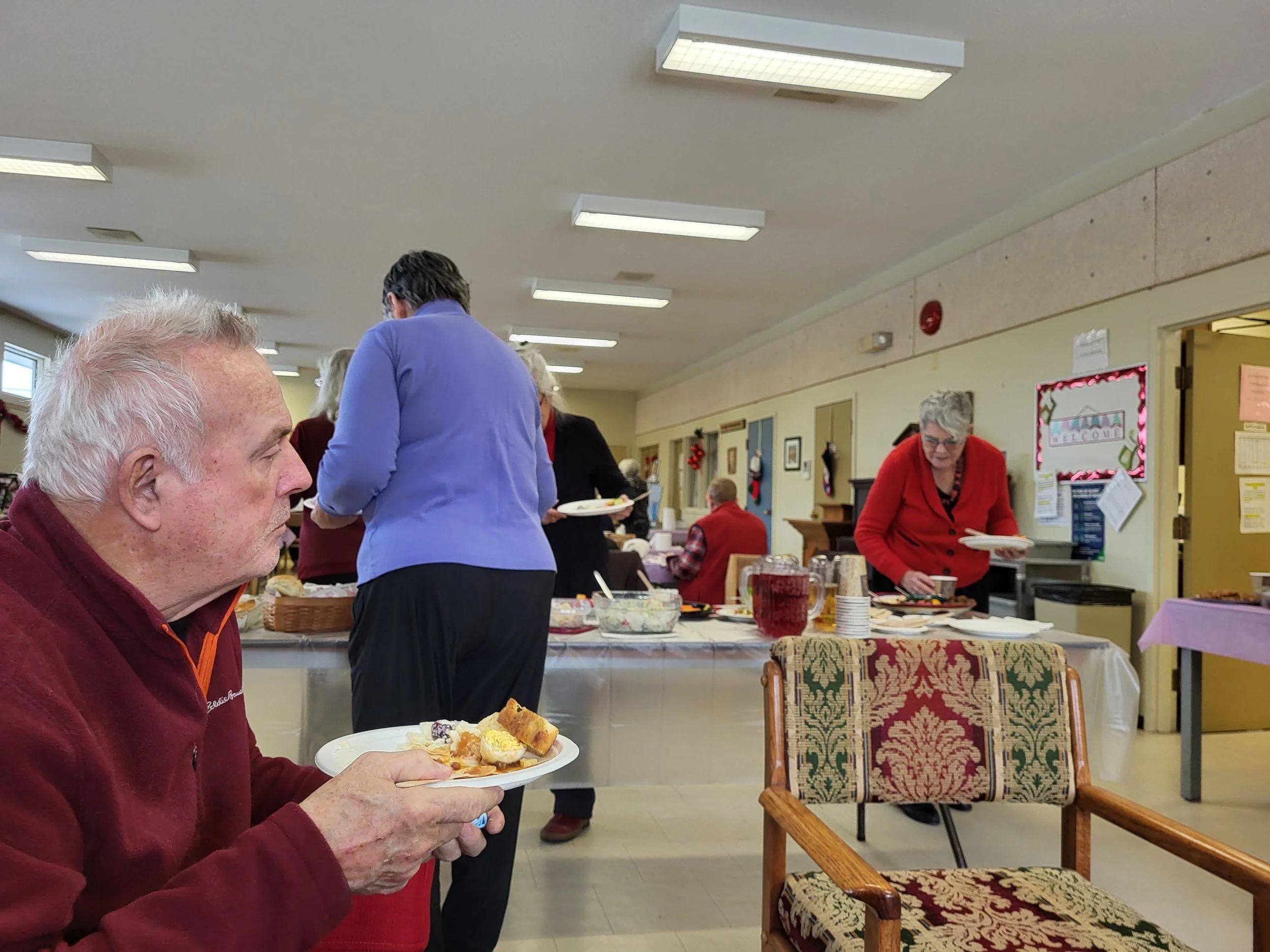 Senior man in red sweater holding a plate of food at a festive buffet in a decorated room.