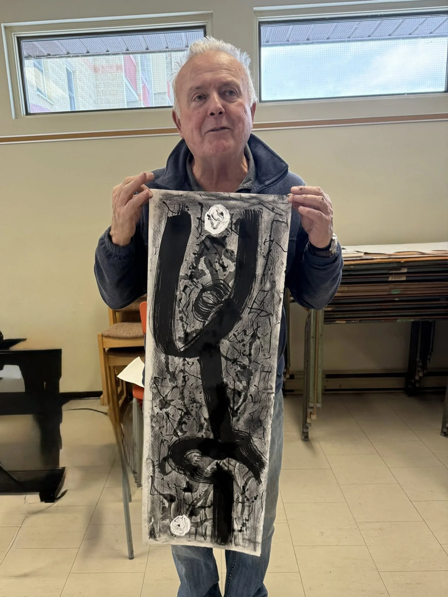 An elderly man with gray hair holding a tall abstract black and white painting, standing in a room with windows in the background.