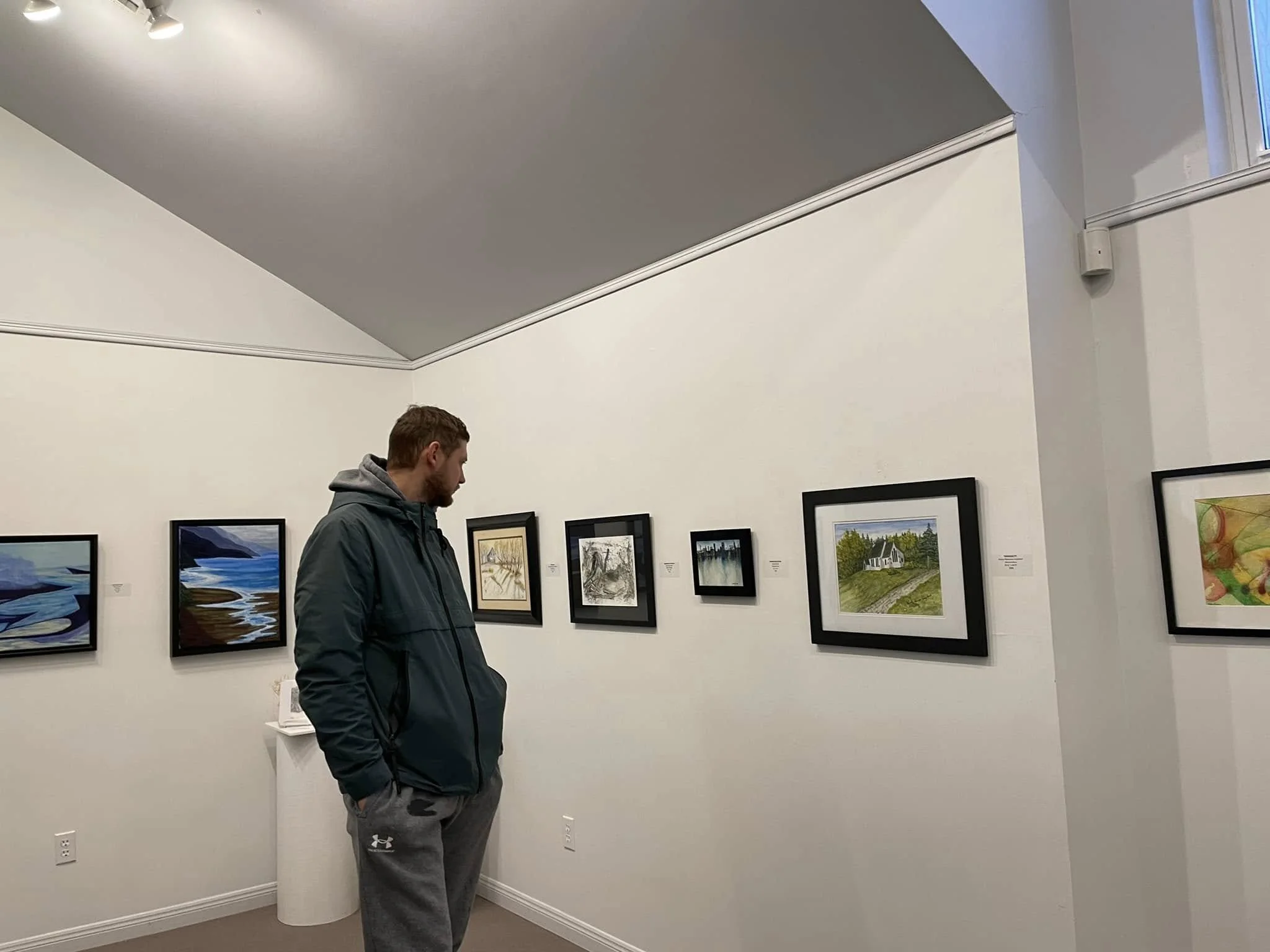A man stands in an art gallery looking at framed paintings on the wall. The walls are white, and there is a small pedestal in the corner.