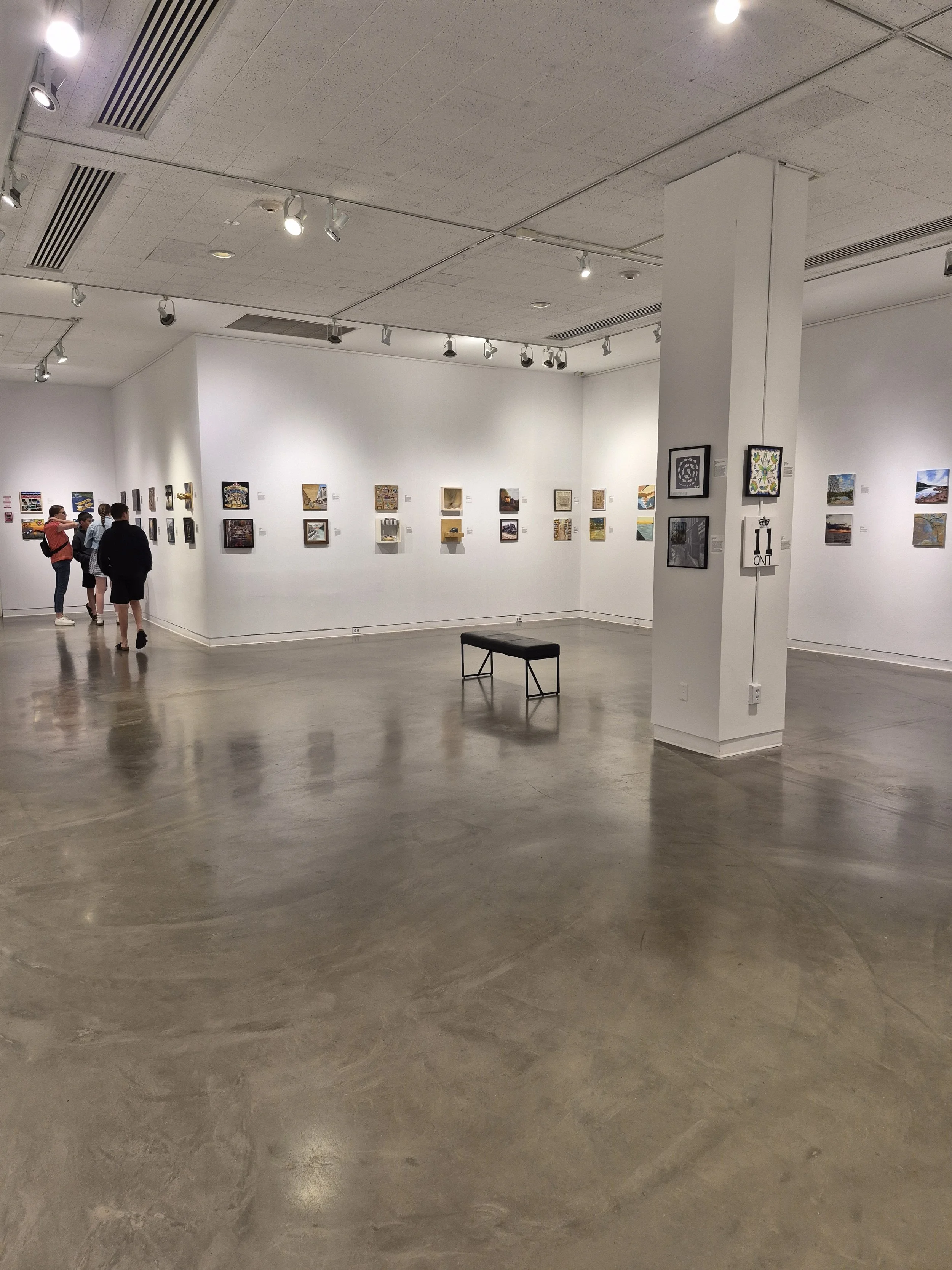 An art gallery with white walls displaying small paintings. Four people are viewing the artwork in the background. There is a bench in the middle of the room, and lighting fixtures are illuminating the paintings.