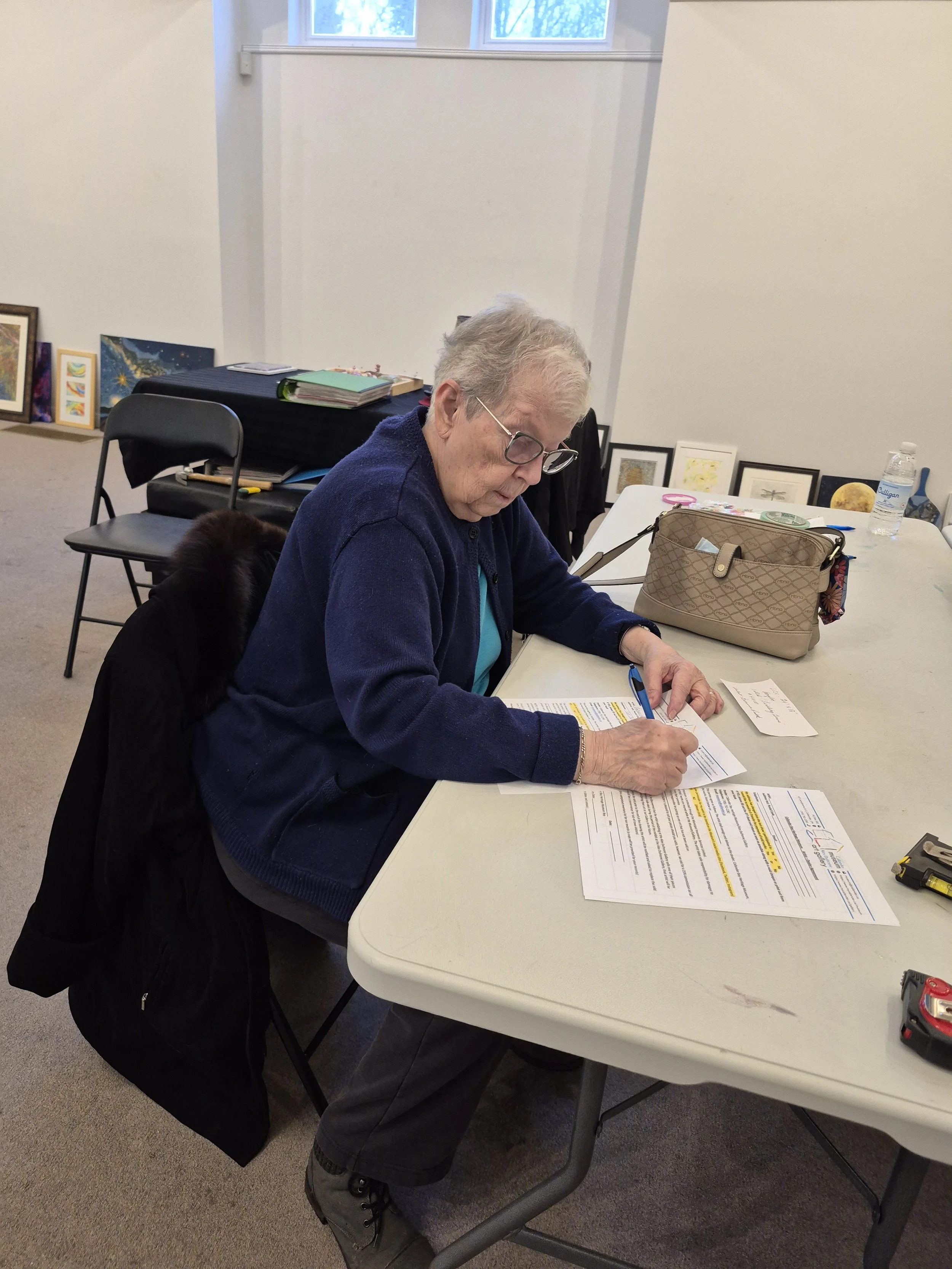 An elderly woman with short gray hair and glasses is sitting at a table, filling out paperwork with a blue pen. She is wearing a navy blue jacket and a turquoise shirt. On the table are additional papers, a tan purse, a water bottle, and some small items. Behind her are framed artworks and a table with storage boxes.