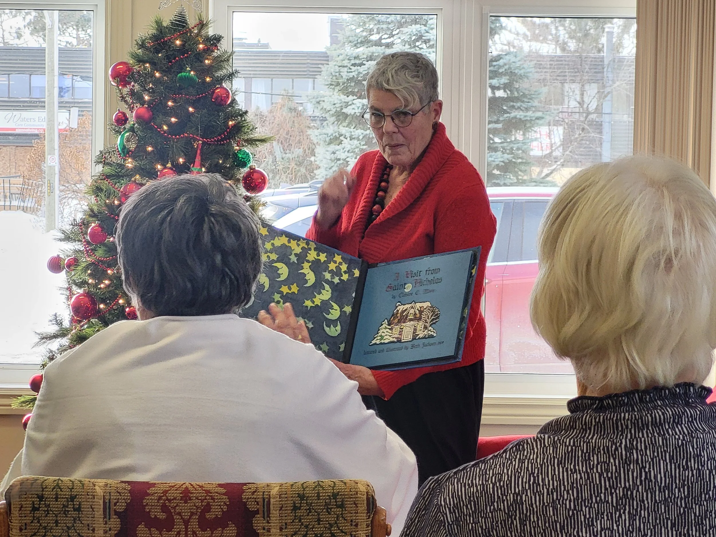 A woman in a red sweater reads a Christmas storybook to two other women seated in front of her near a decorated Christmas tree with red and green ornaments. Outside the window, snow and parked cars are visible.