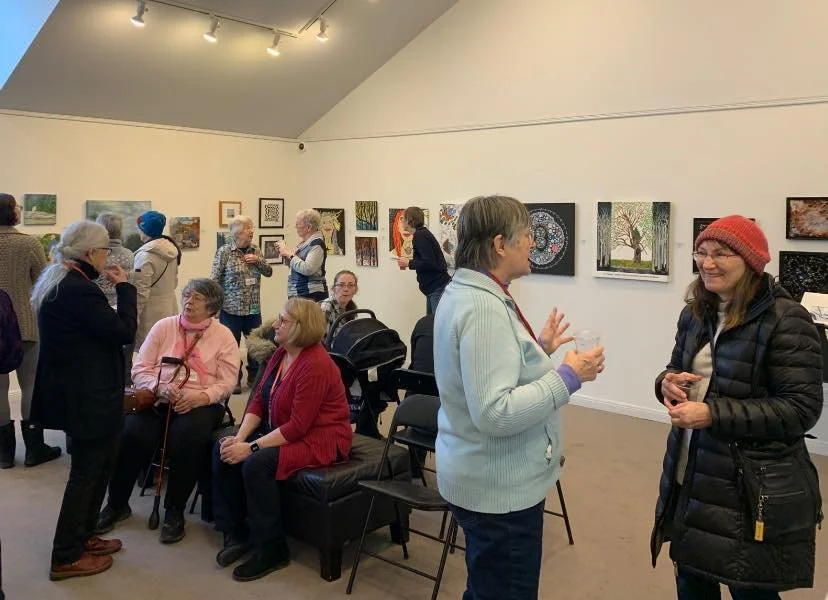 People mingling and chatting in an art gallery with various paintings hanging on the white walls.