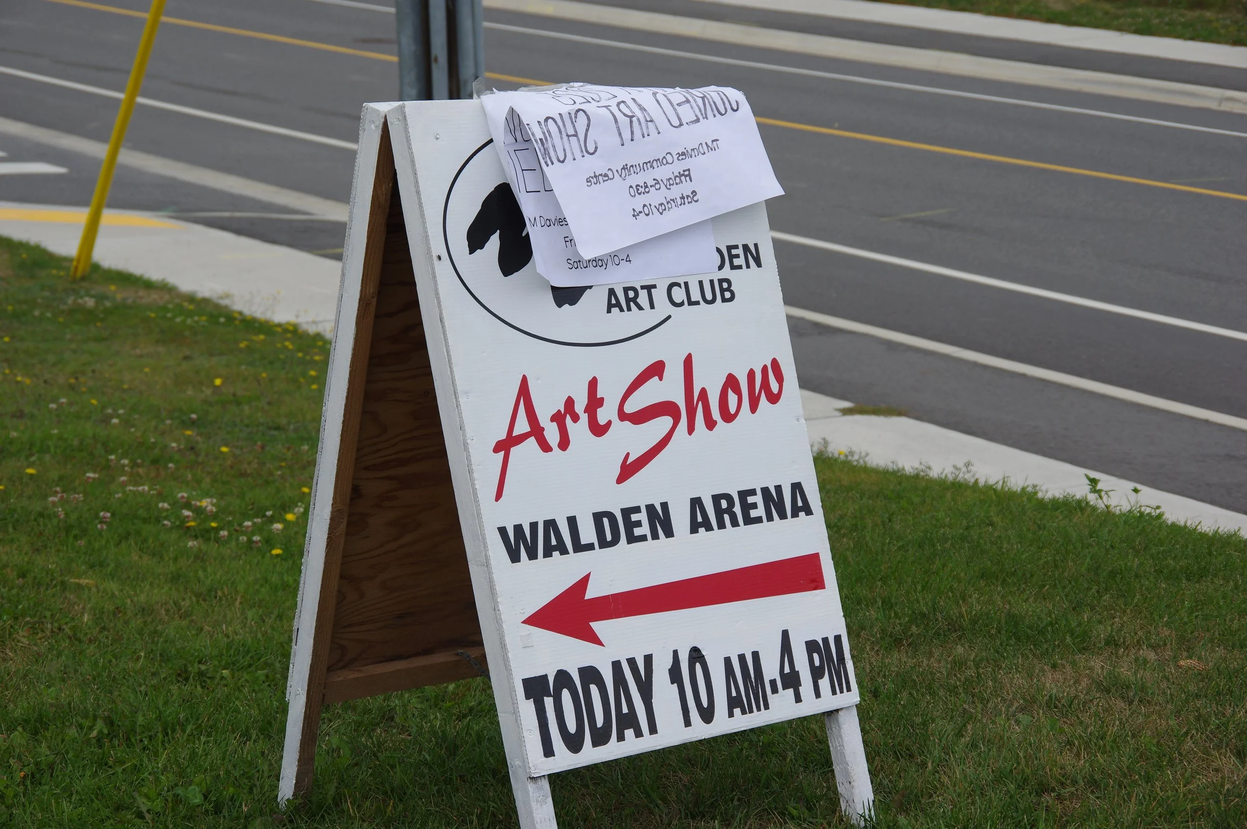 A sidewalk sign outside Walden Arena directing to an art show at Walden Art Club from 10 a.m. to 4 p.m. with an arrow pointing left. A second paper sign is attached to the top of the sign, partially covering the main sign.