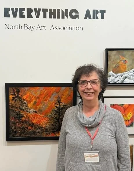 A woman with glasses and a gray sweater standing in front of artwork at an art exhibition. The wall behind her has the words "EVERYTHING ART" and "North Bay Art Association" written on it, with framed paintings visible.