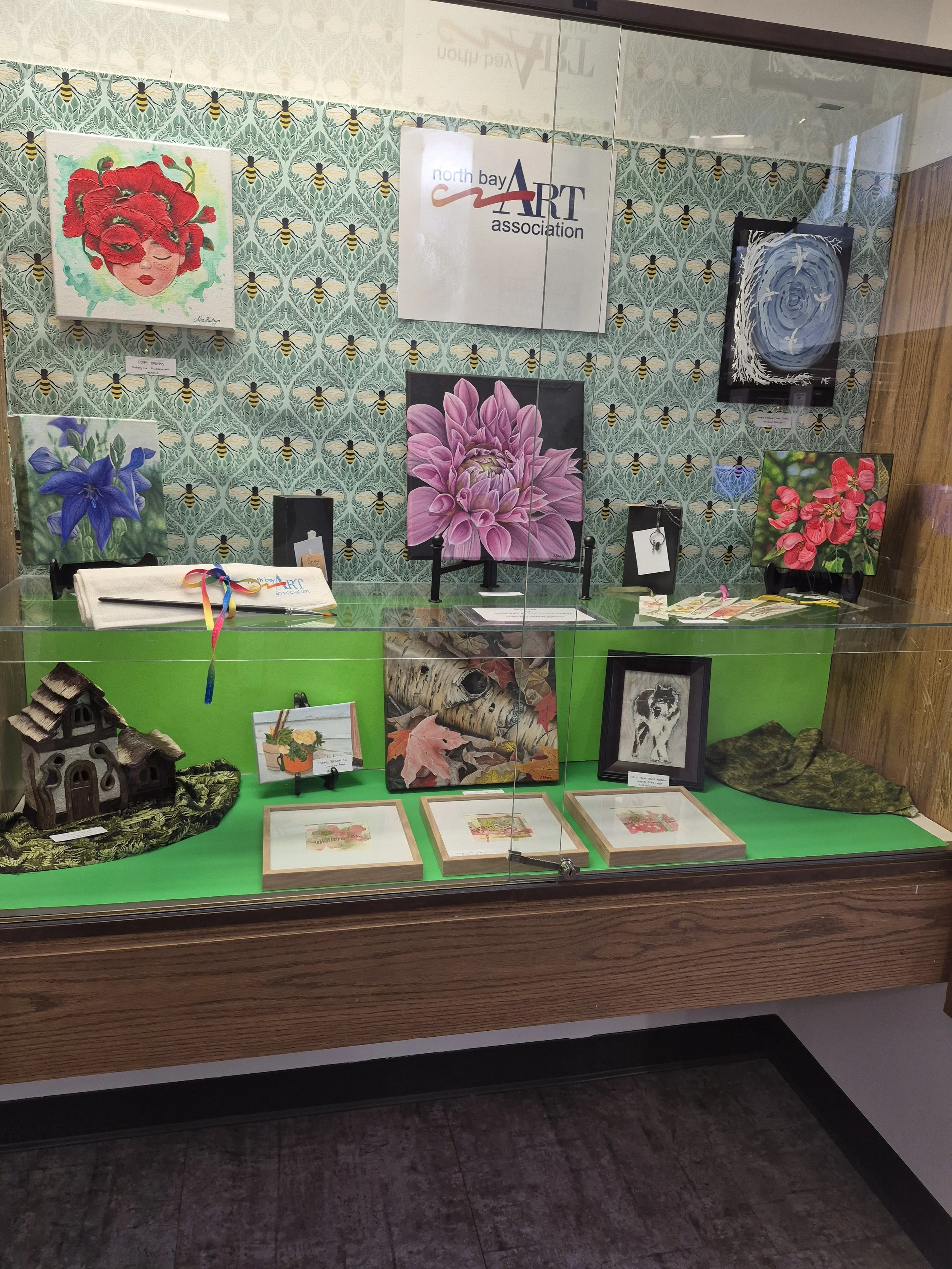 A glass display case showcasing various artwork and crafts, including floral paintings, miniature framed paintings, and small decorative items, with an art association sign in the background.