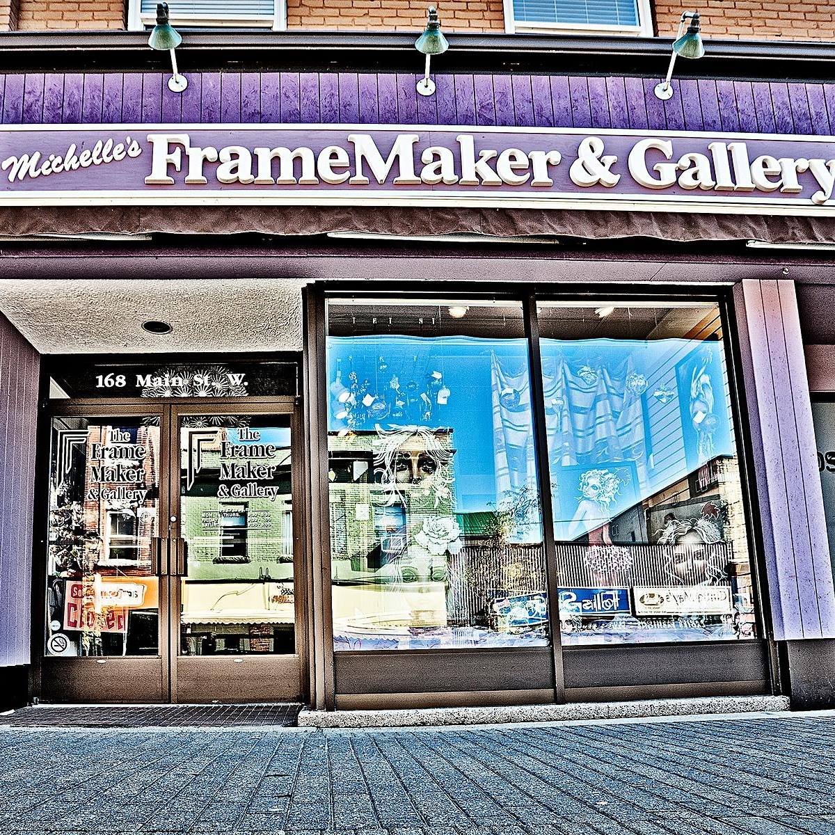 storefront of Michelle's FrameMaker & Gallery with glass doors and windows, reflecting street scene, decorative paintings and figures inside, purple sign with white lettering, brick building above, located at 168 Main Street W.