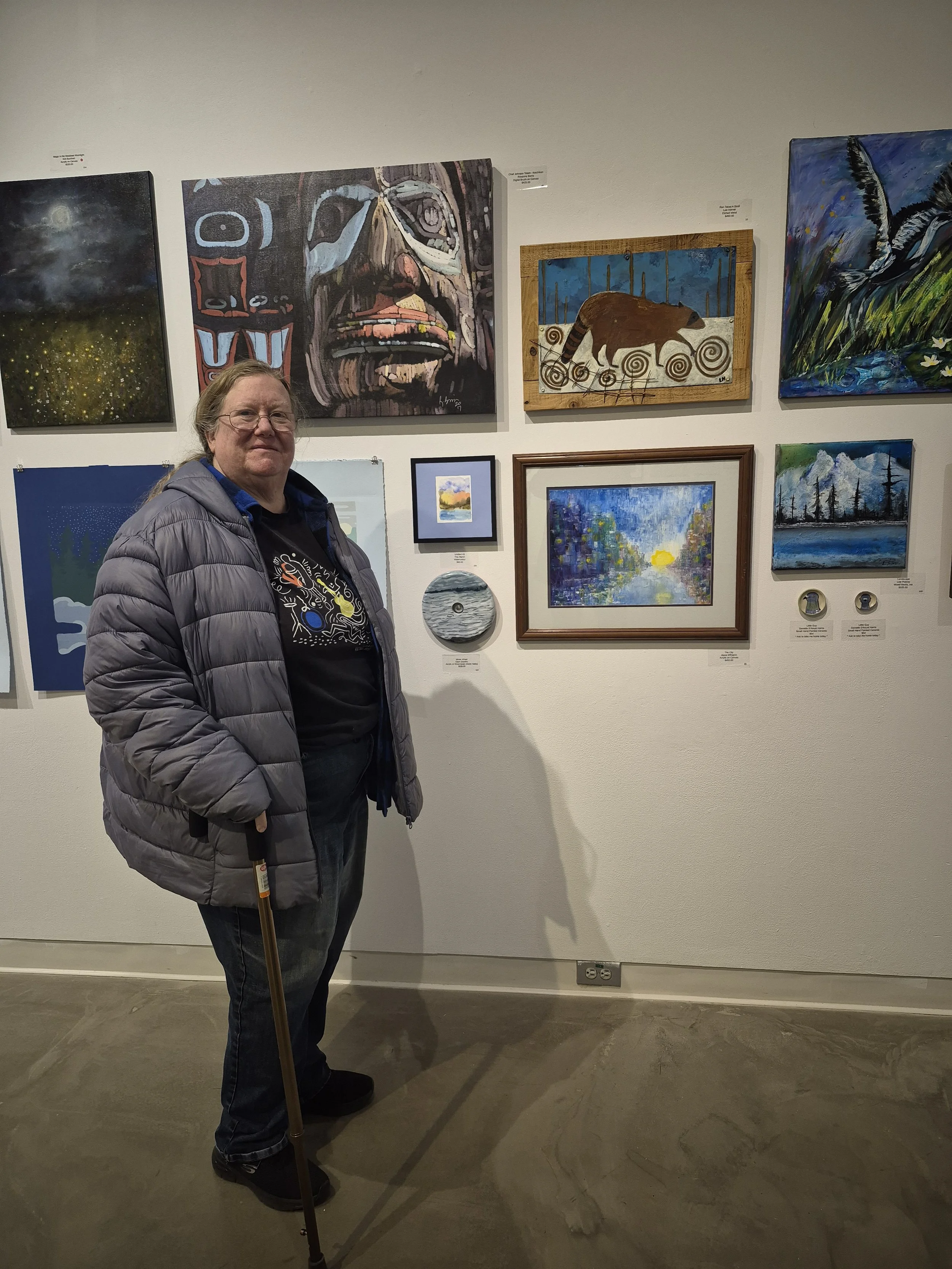 A woman with glasses and a cane standing in front of a wall of colorful paintings in an art gallery.