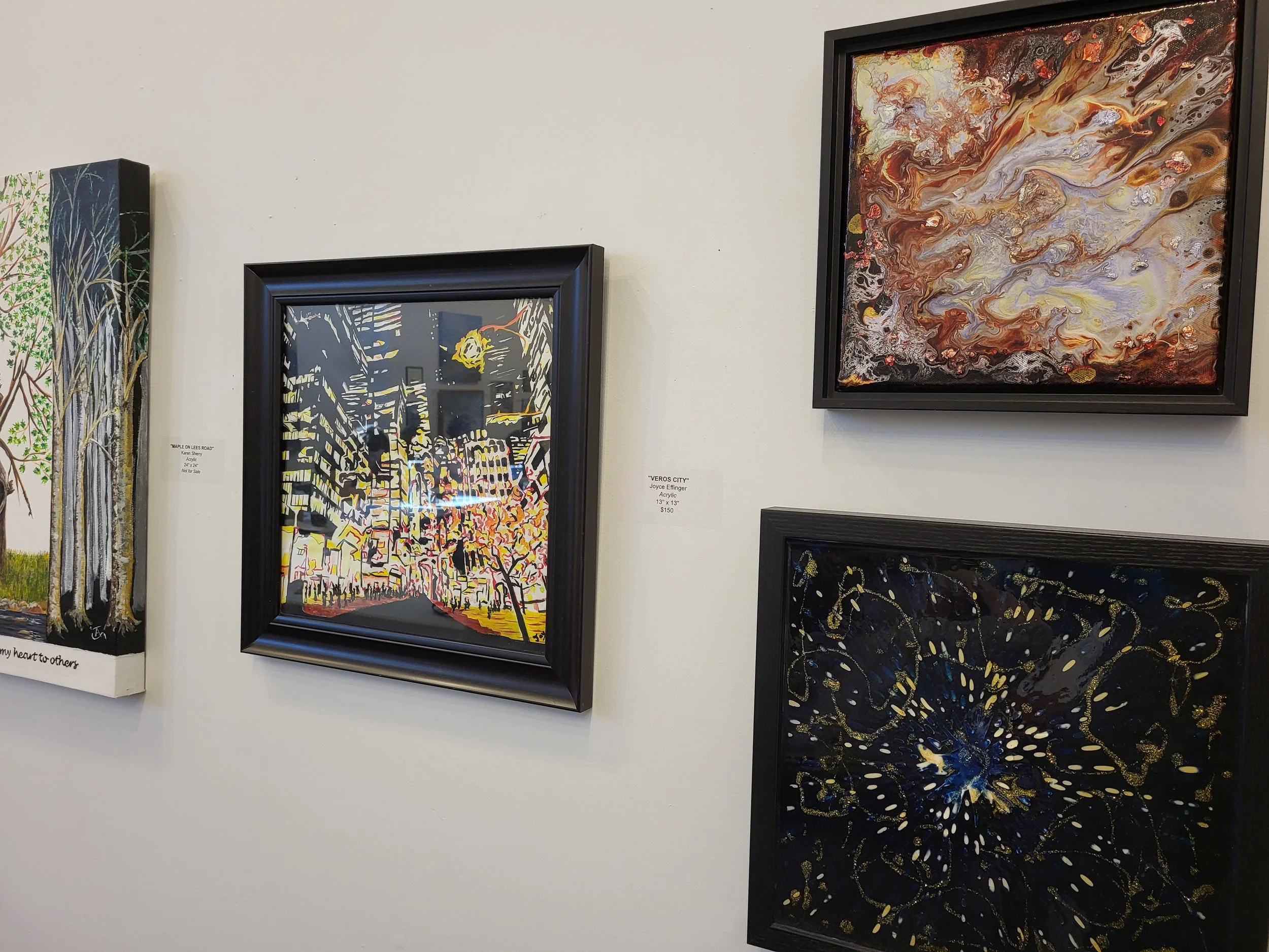Gallery wall with four framed art pieces, including cityscape, abstract explosion with flowing colors, colorful butterfly, and a tree scene, in a museum or gallery setting.