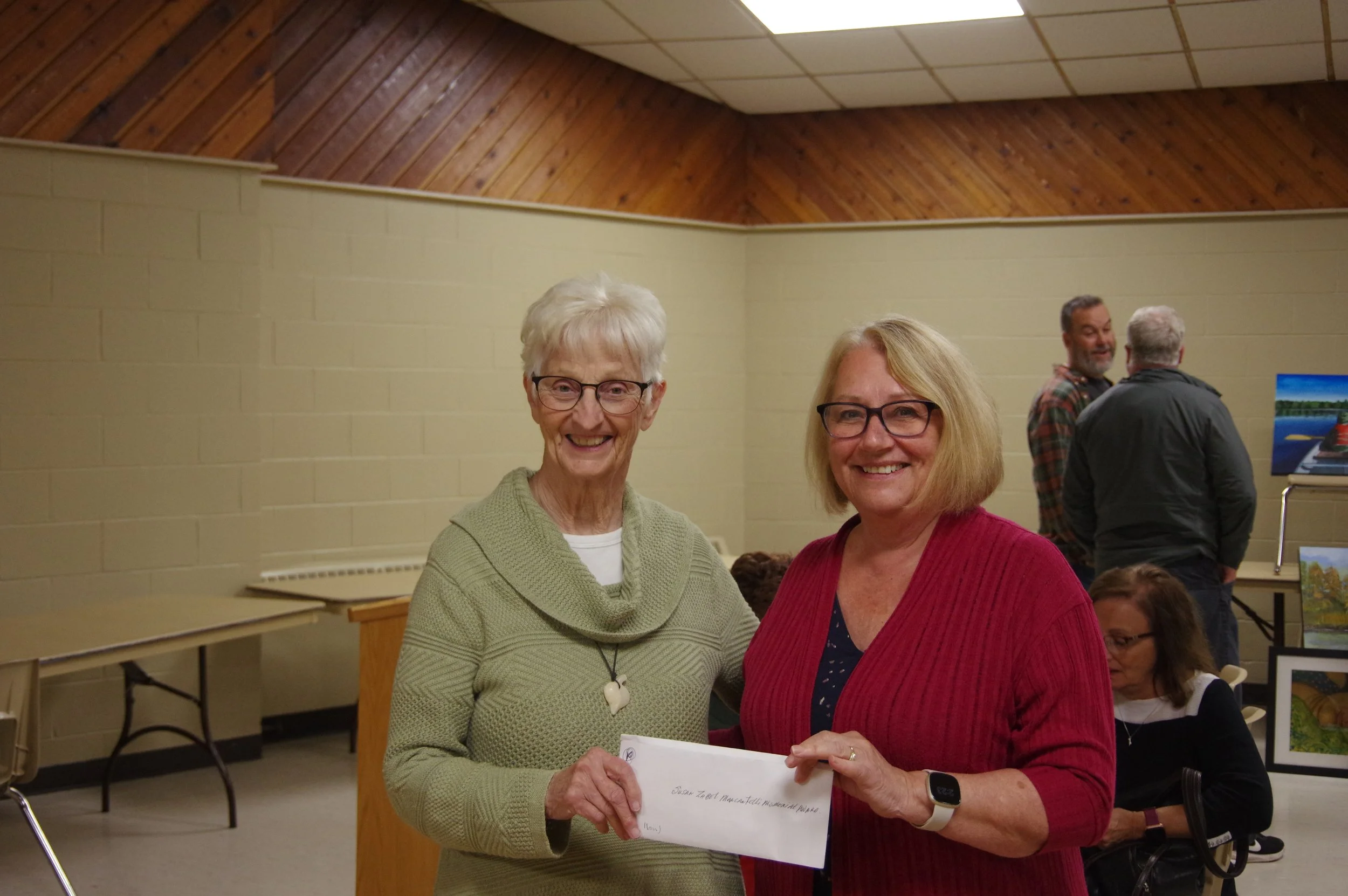 Two women smiling and holding an envelope at an indoor event, with other people in the background.