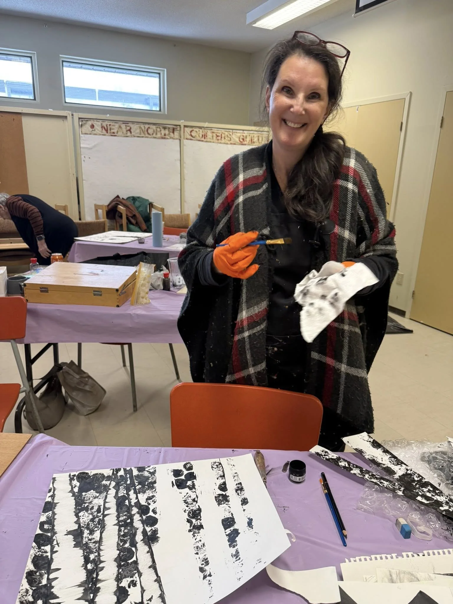 A smiling woman with glasses on her head wearing an orange glove, black shirt, and plaid shawl, holding a paintbrush and paper towel, in an art studio with tables covered in purple cloth and art supplies, creating black and white artwork.