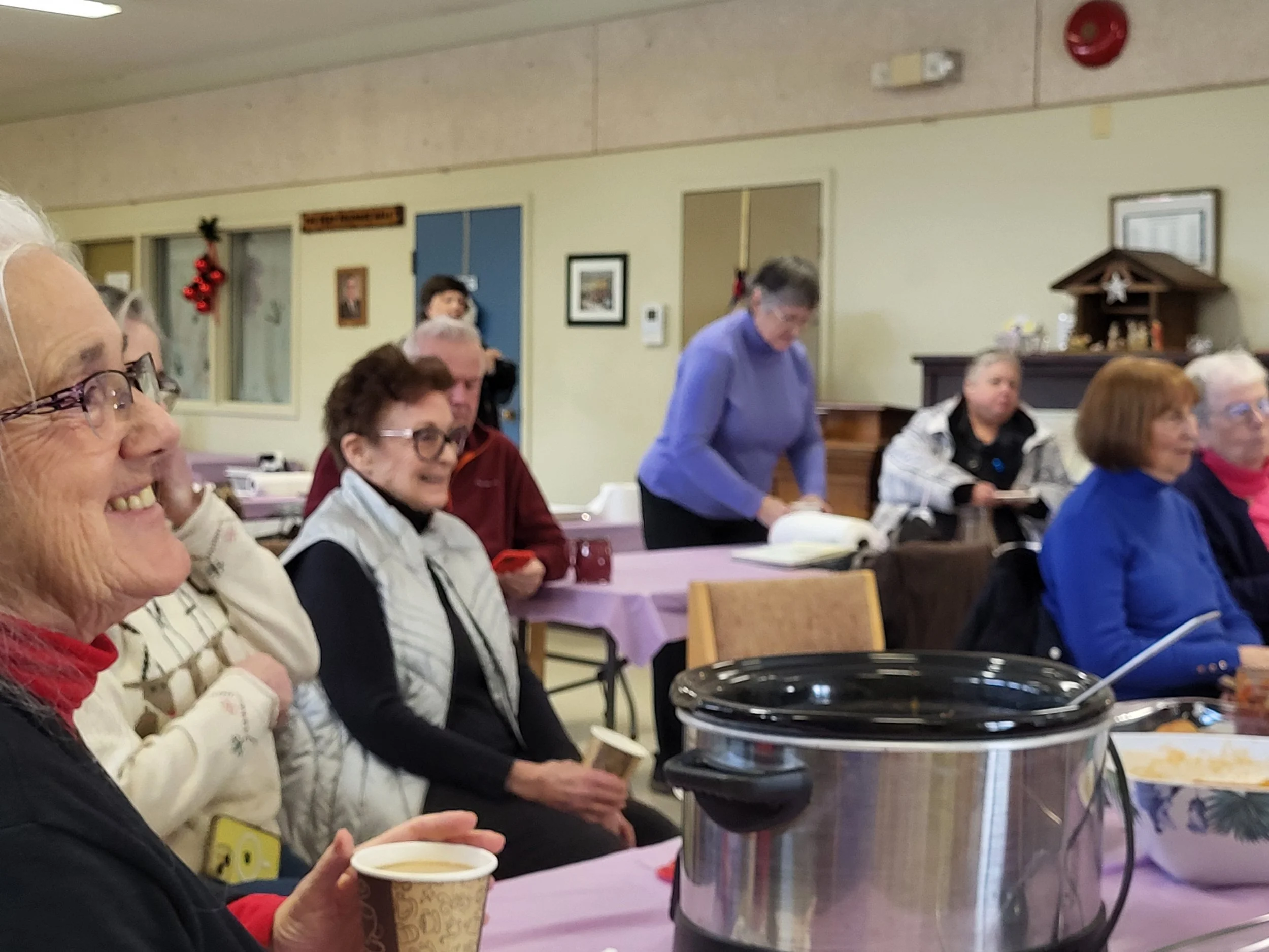 Group of elderly people sitting at tables during a gathering, smiling and enjoying food and drinks, with some serving themselves from a large slow cooker, in a decorated community room.