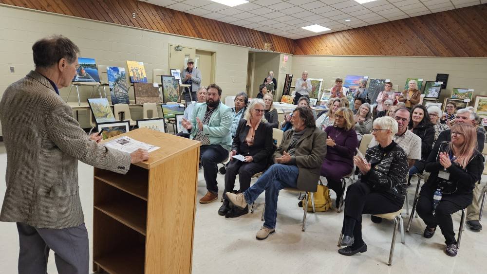 A man giving a presentation or speech to a seated audience at an art exhibit or gallery. Paintings are displayed on tables and easels in the background.