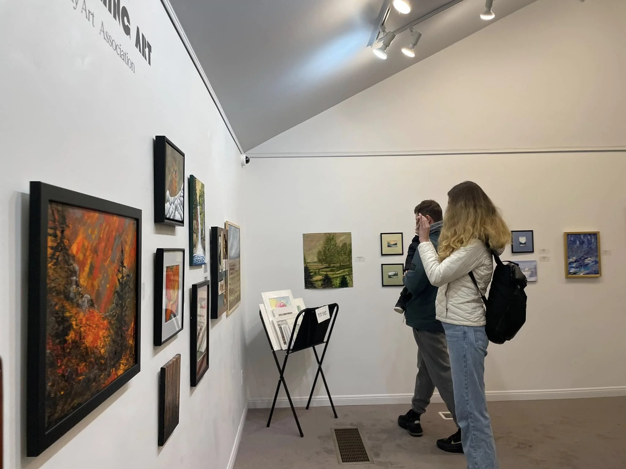 Two people at an art gallery, one woman with a backpack examining paintings on the wall, while a man stands nearby looking at artwork.