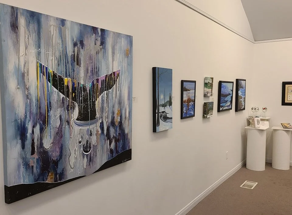 An art gallery wall displays various paintings. The largest painting features a whale with colorful dripping paint. Smaller landscape paintings depict snowy scenes, rivers, and lakes. There are two white pedestal tables with decorative items on them.