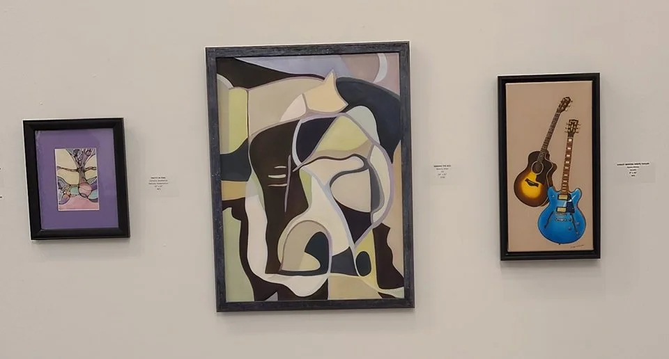 Three framed artworks on a beige wall, including an abstract painting in the center, a stylized butterfly drawing on the left, and two guitars, one yellow and one blue, on the right.