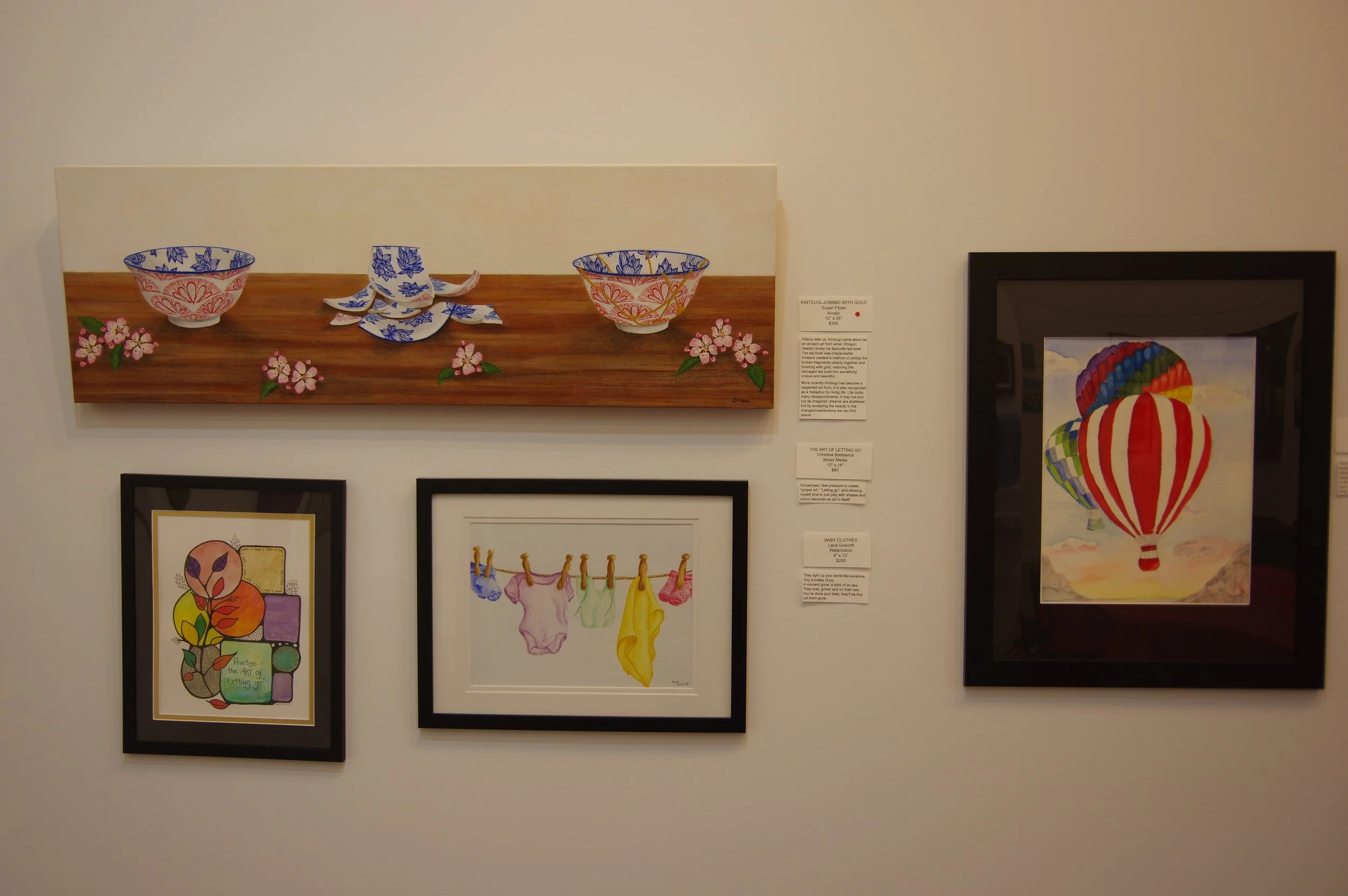 Art display of watercolor paintings featuring colorful bowls, fruit, hanging laundry, hot air balloons, and floral motifs on a white gallery wall.