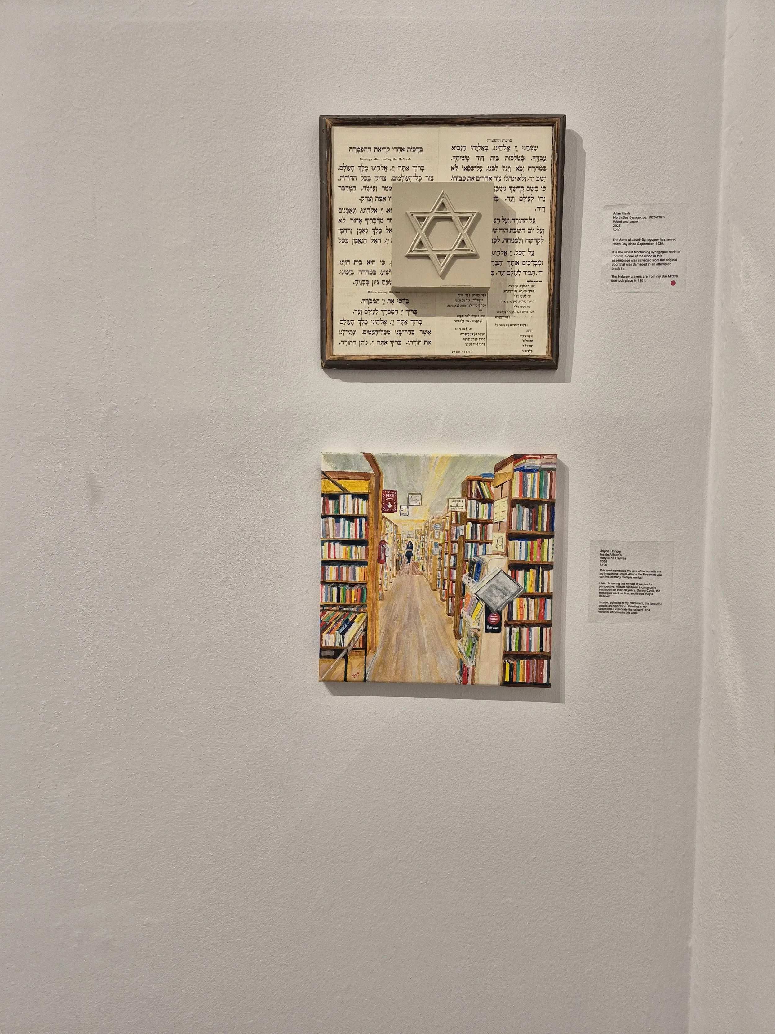 Image of two artworks on a white gallery wall. The top artwork features a framed Hebrew text with a Star of David symbol in the center. The bottom artwork is a colorful painting of a library aisle filled with books.