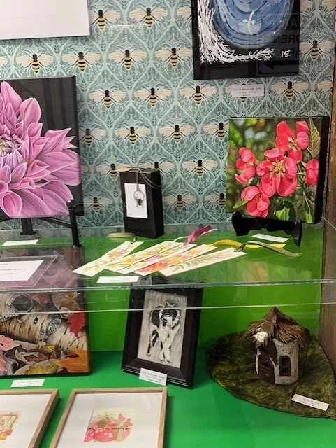Display of artwork and decorative items, including floral paintings, a cat-themed framed picture, and a small wooden house sculpture, arranged on a green and transparent table.