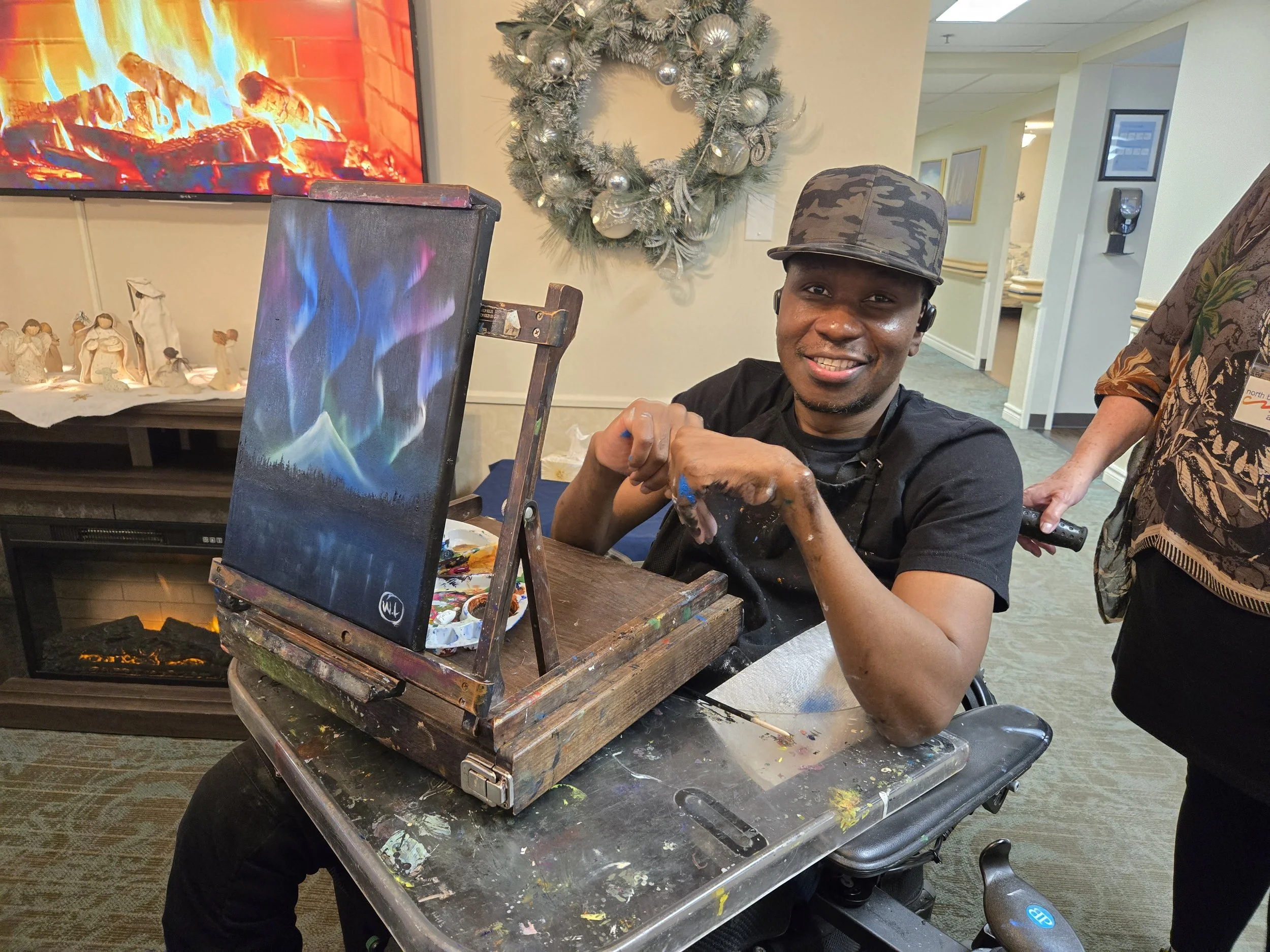 A man in a wheelchair smiling at the camera while painting a picture of the northern lights, with Christmas decorations and a fireplace visible in the background.