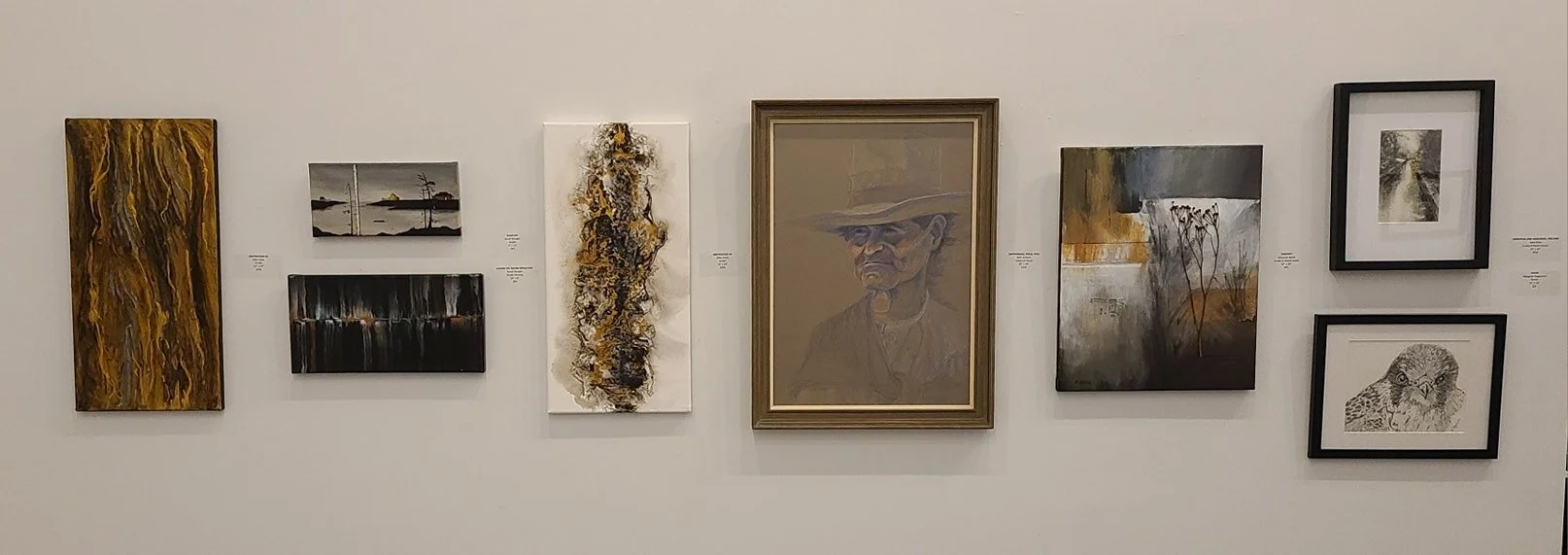 Gallery wall with seven framed artworks: abstract paintings, landscape, portrait of a man with a hat, a sketch of a bird, and other abstract and landscape art.