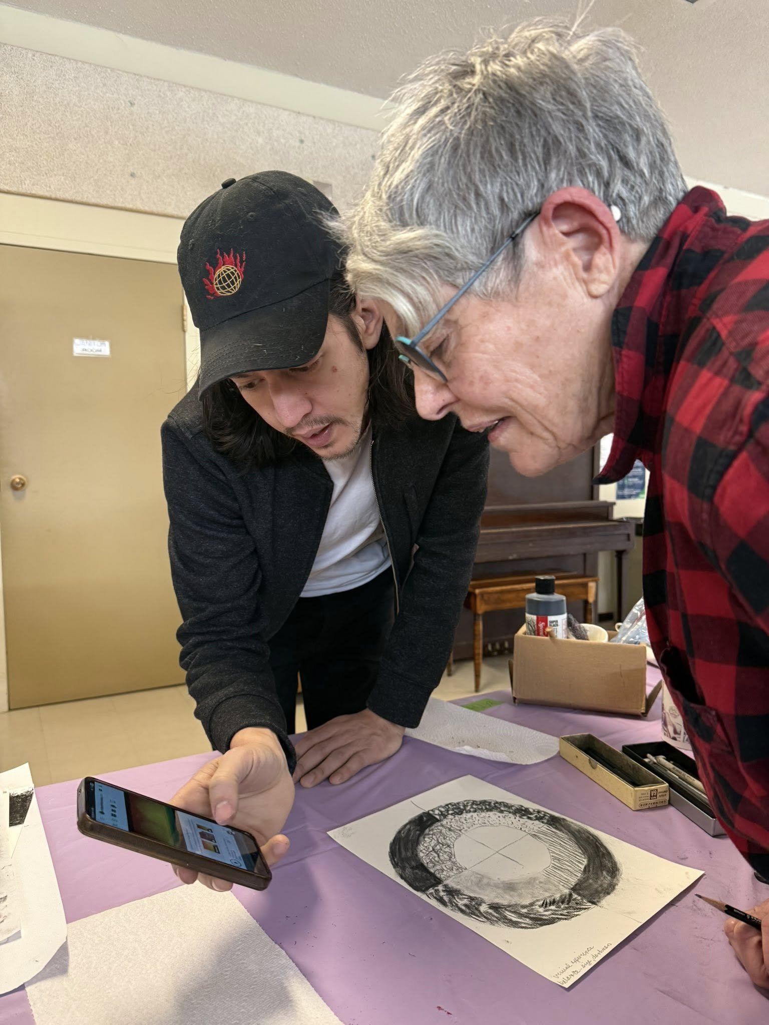 Two people, an older woman and a young man, lean over a table looking at a smartphone, with artwork on the table in front of them.