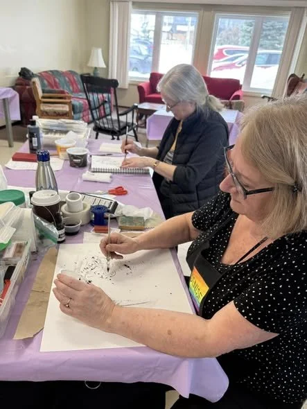 Two women working on creative projects at a table covered with craft supplies in a cozy, well-lit room with large windows and comfortable chairs in the background.