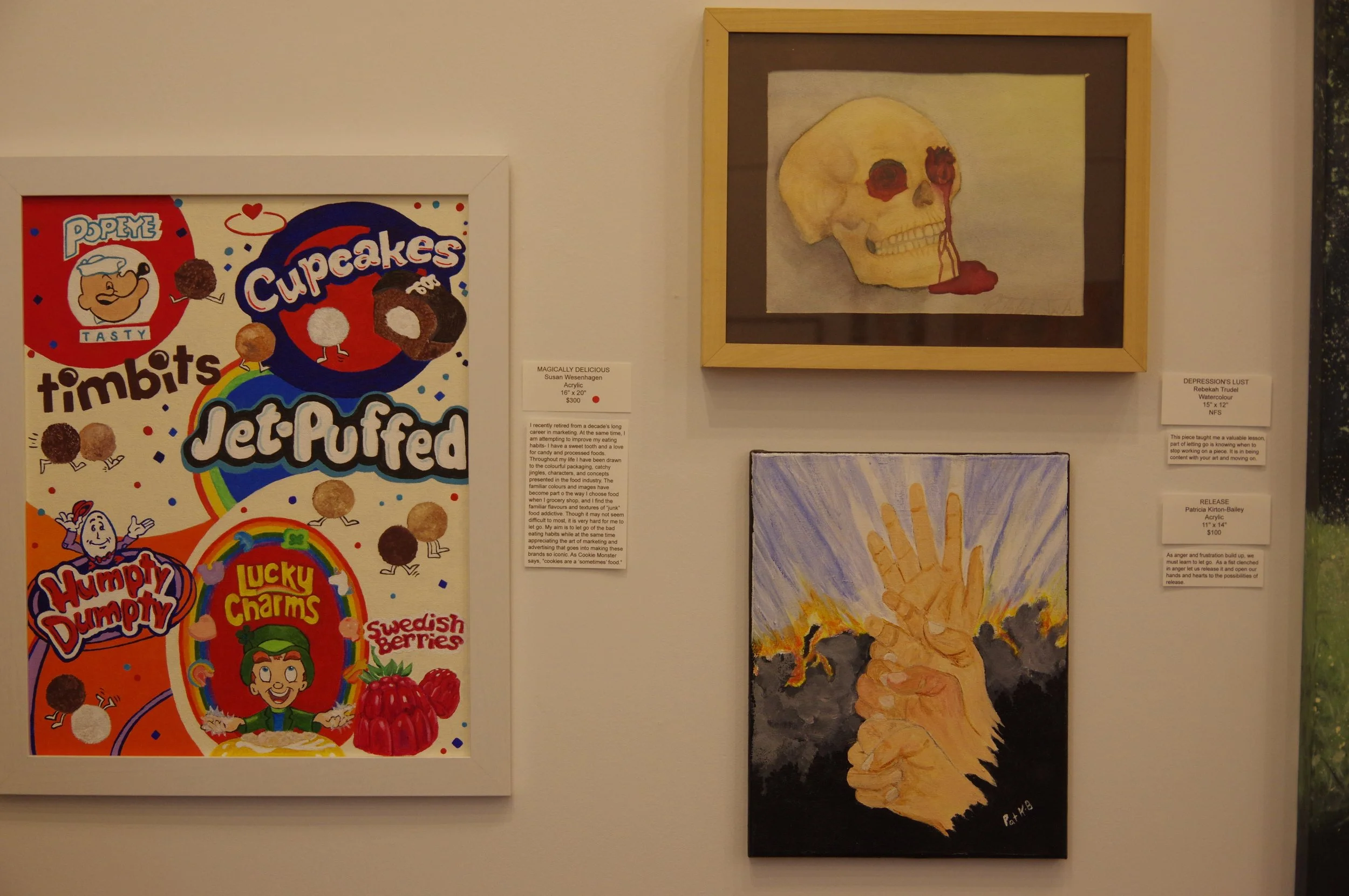 Photograph of three framed artworks on a gallery wall. The large colorful artwork on the left features cartoon illustrations of various snacks like cupcakes and Swedish berries with playful text, titled 'MAGICALLY DELICIOUS' by Susan Wiesenhagen. The top right artwork is a watercolor painting of a human skull with red accents, titled 'DEPRESSION'S LUST' by Rebekah Trudel. The bottom right artwork depicts a hand with extended fingers, surrounded by lightning, titled 'RELEASE' by Patricia Kenton-Bailey.