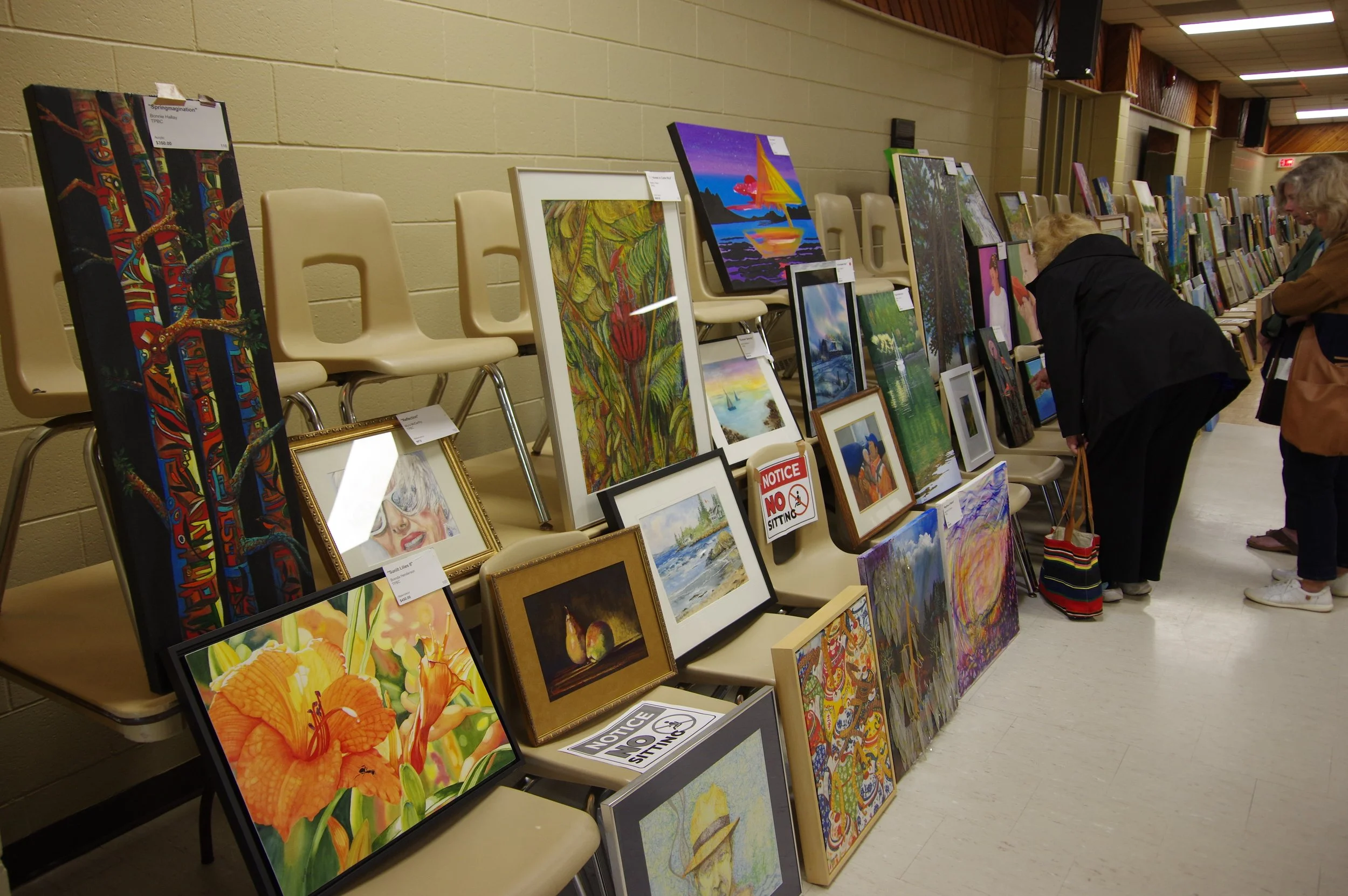 An art display with various paintings and framed pictures arranged on chairs and the floor, with people browsing the artwork in a well-lit indoor space.