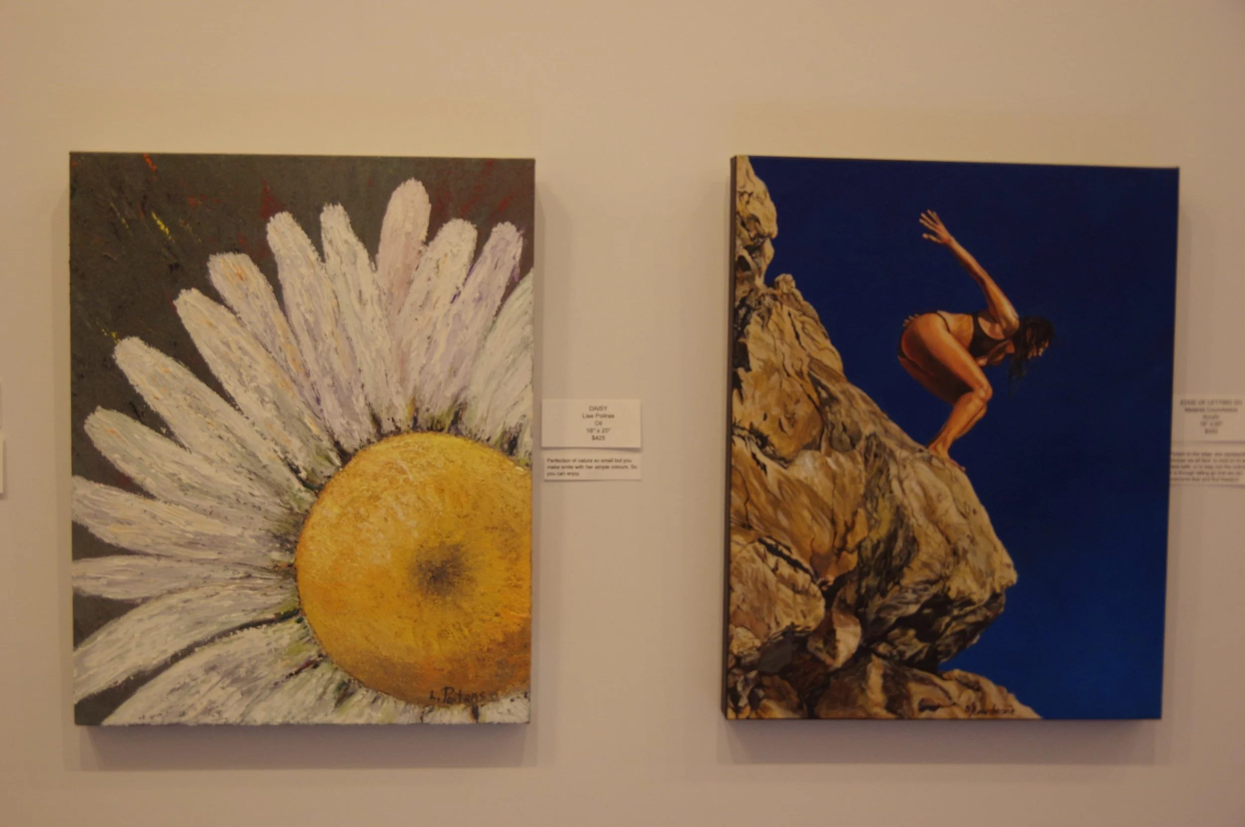 Two paintings hang on a gallery wall; the left painting is an impressionist-style of a large daisy flower with white petals and a yellow center, and the right painting depicts a woman in a swimsuit crouching on rocks against a dark blue sky.