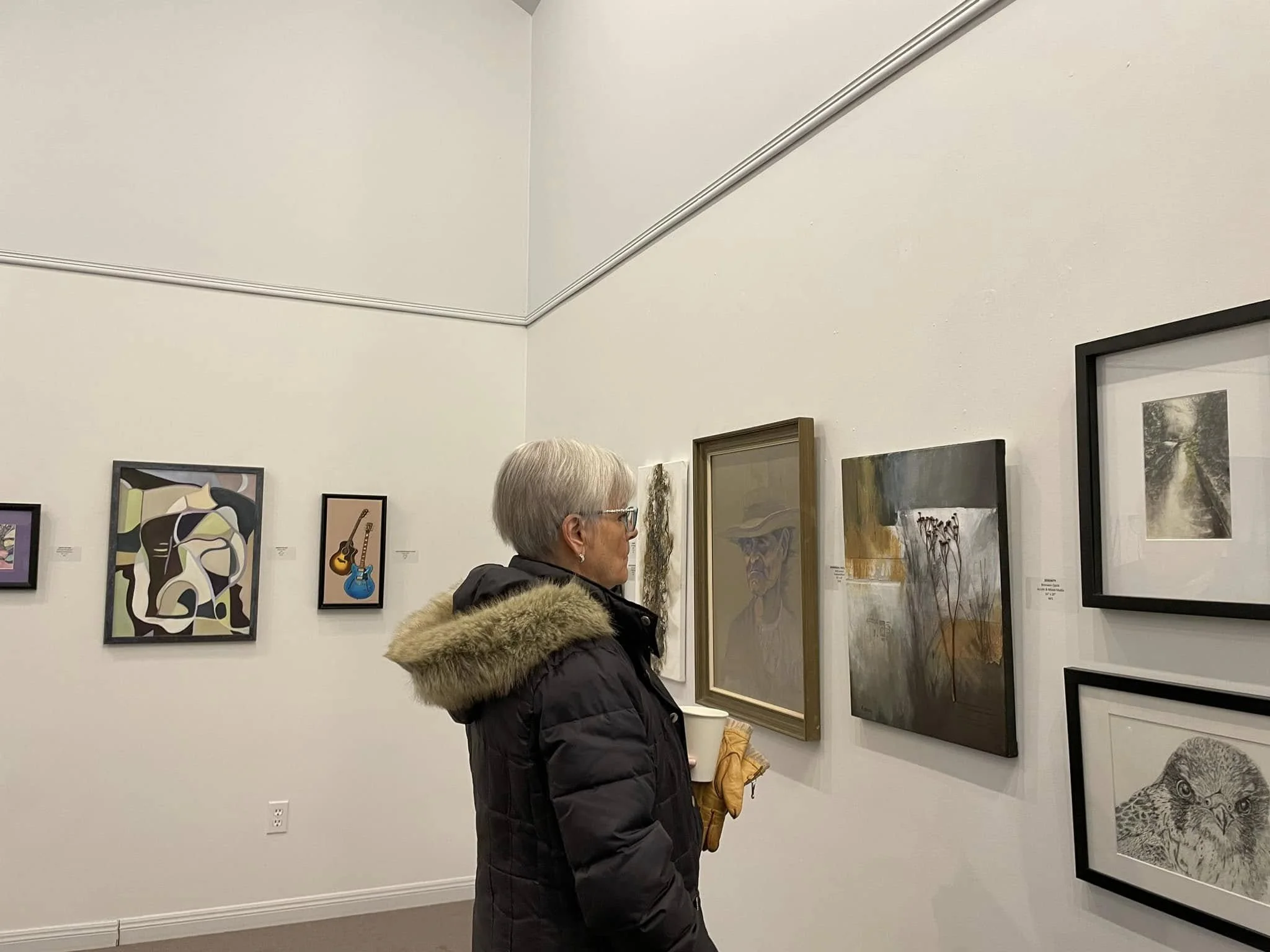 A woman with gray hair and glasses stands in an art gallery, observing various paintings on the wall, including abstract, realistic, and black-and-white artwork.