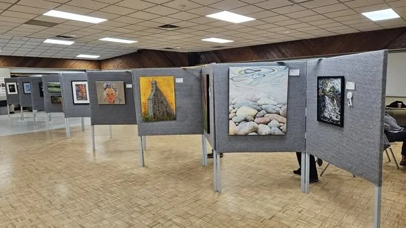 Art exhibition with paintings displayed on gray fabric-covered panels in a spacious room with a wooden floor and ceiling lights.