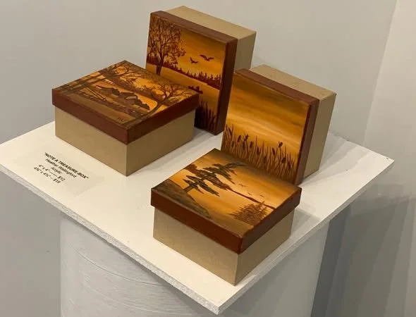 Four wooden boxes with landscape paintings, including a sunset with trees, lake, and birds, displayed on a white pedestal.
