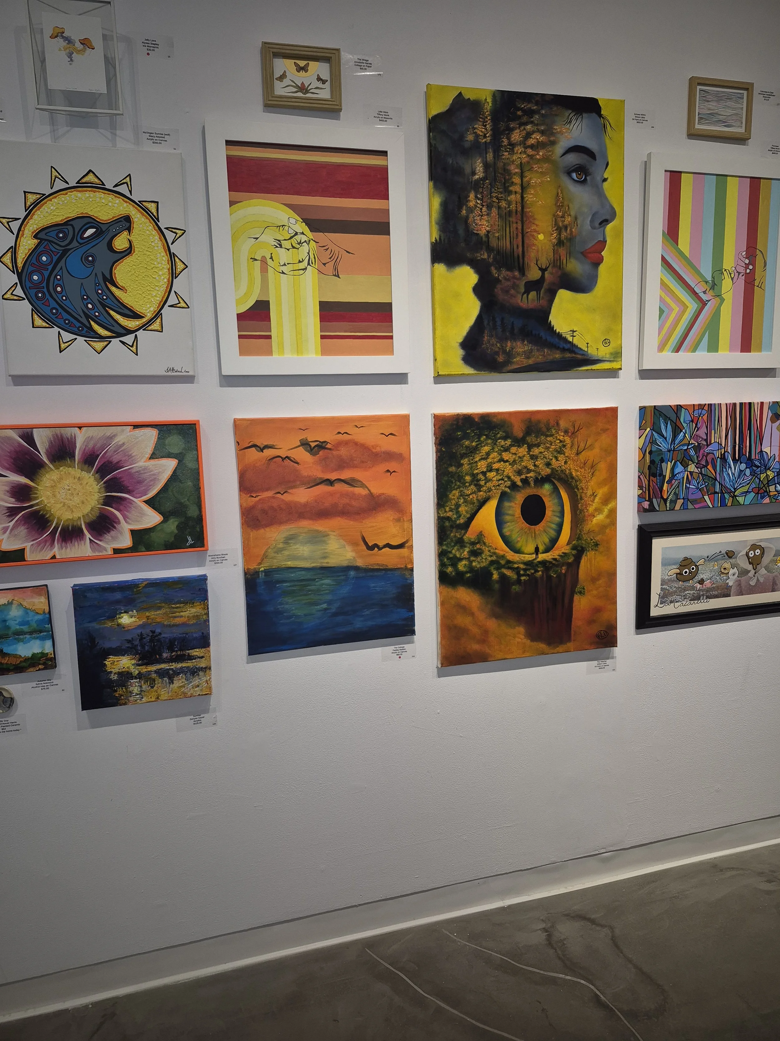 A wall displaying various colorful artworks, including abstract, nature, and surreal paintings, with some framed and others on canvas.