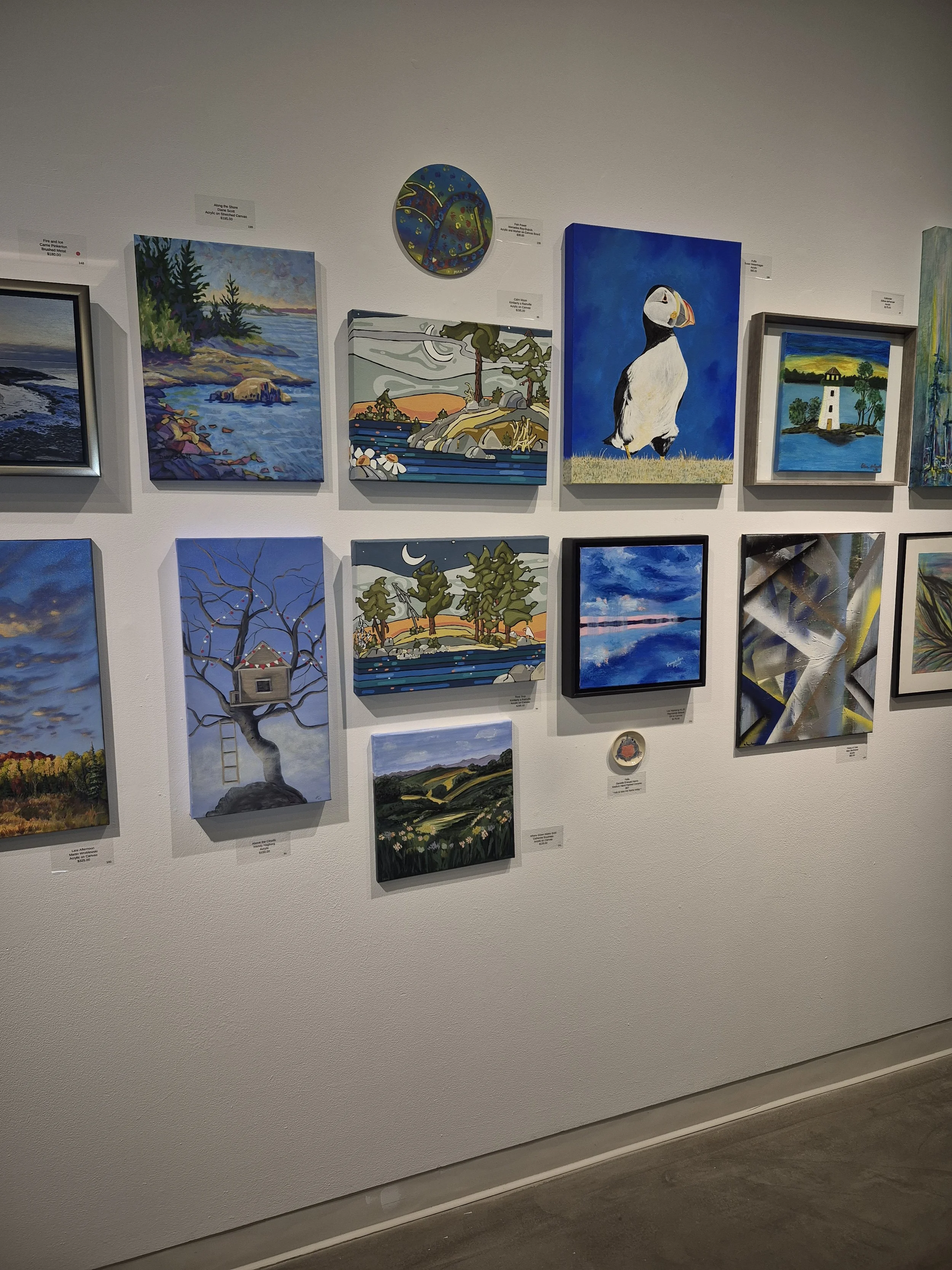 Gallery wall displaying various colorful landscape paintings and a painting of a puffin bird on a blue background.