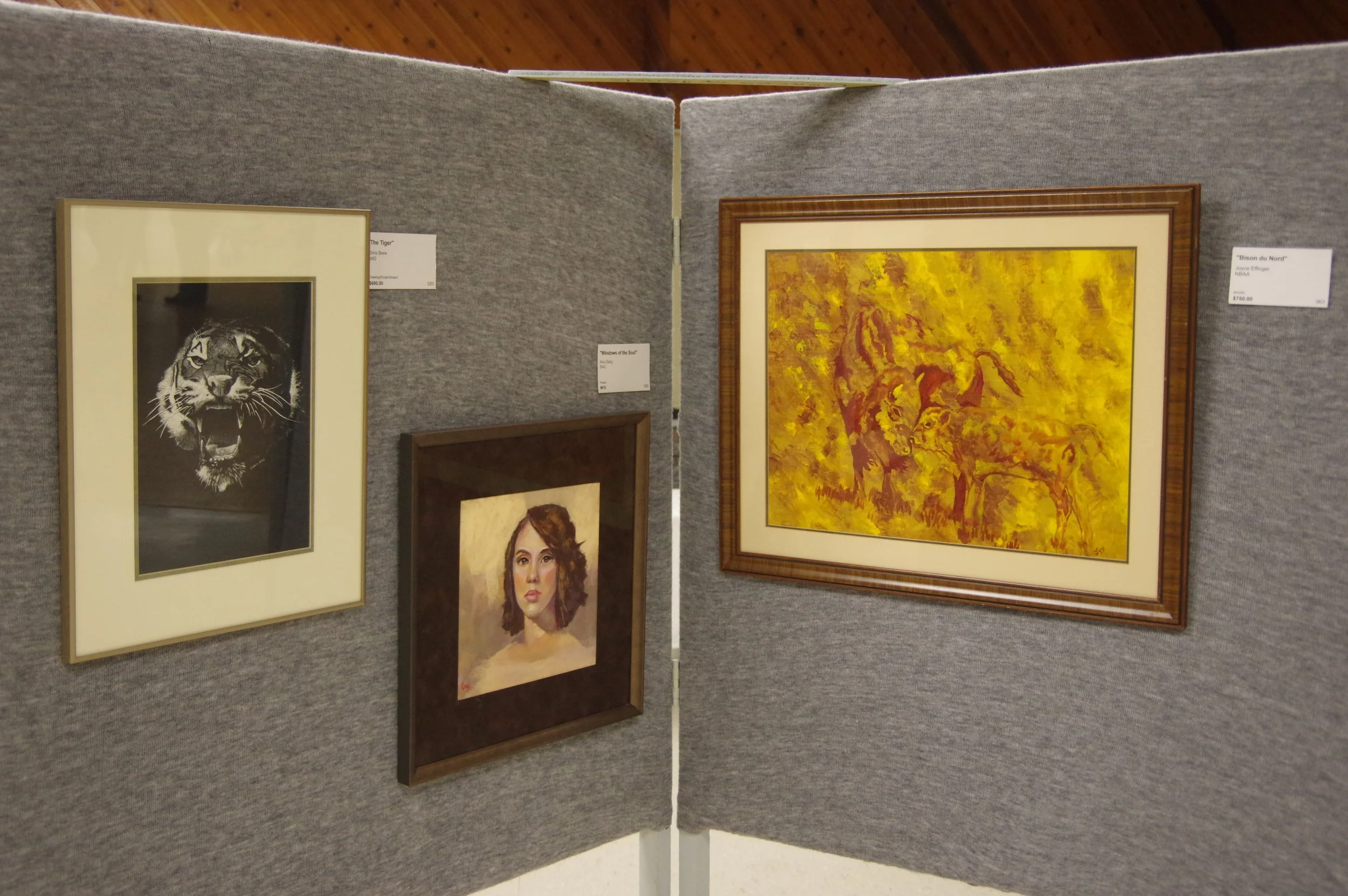 Art gallery display with four framed artworks on gray fabric partition walls, including a black-and-white tiger, a woman's portrait, a colorful warm scene with buffalo, and a landscape.