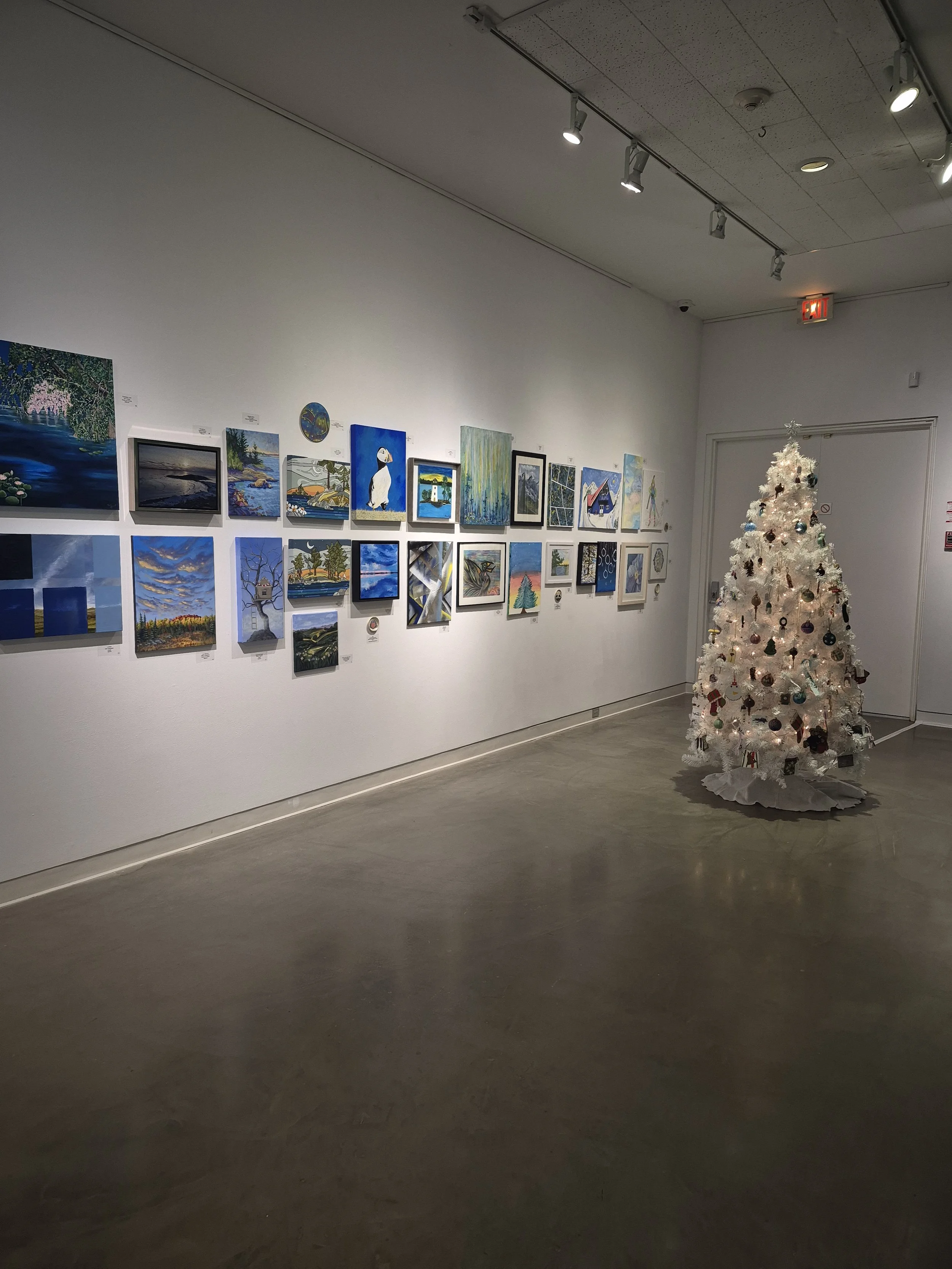 An art gallery with a white wall displaying multiple landscape and figurative paintings. There is a white Christmas tree decorated with ornaments in the corner.