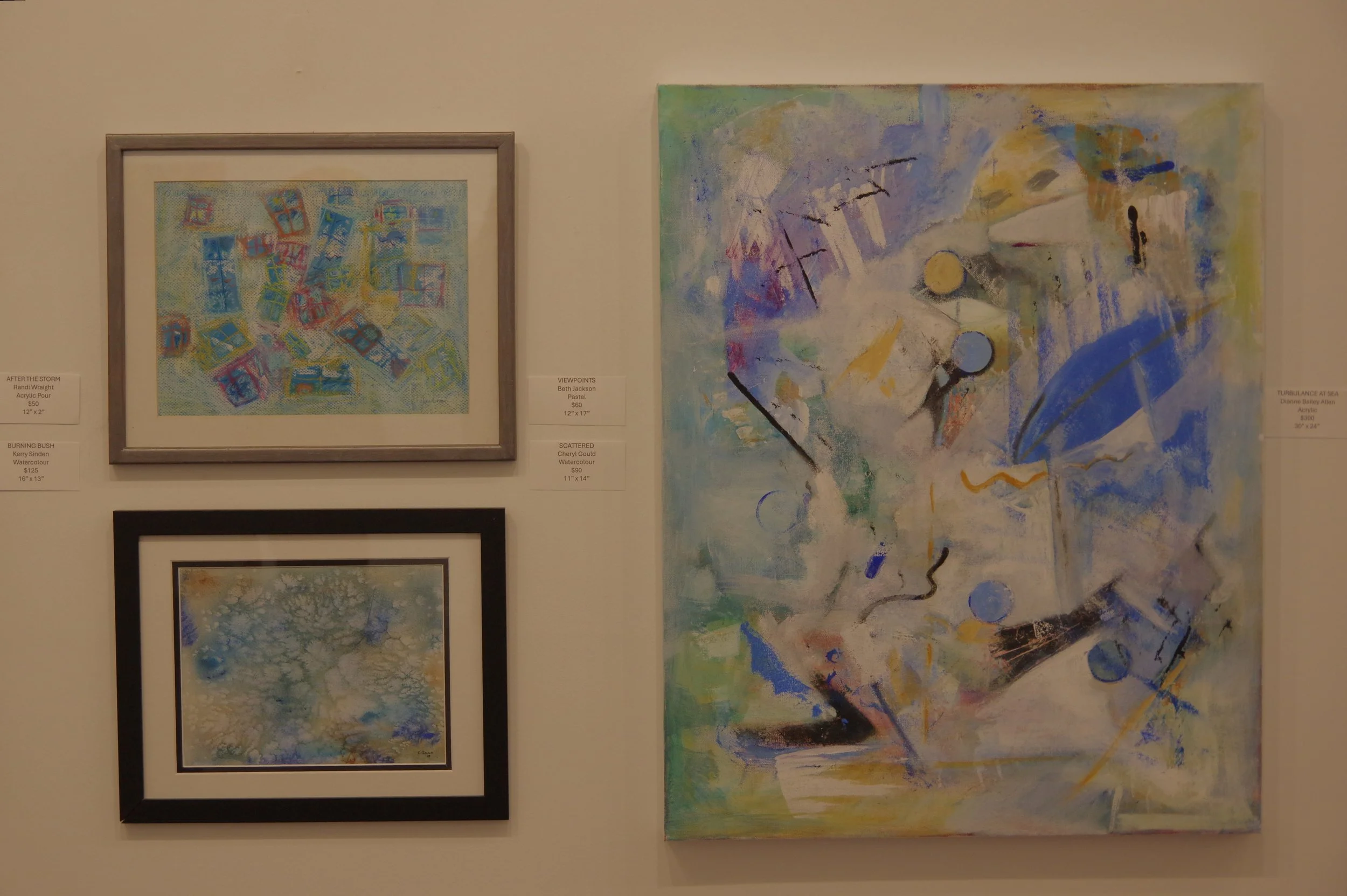 Three abstract paintings hung on a beige wall, featuring geometric and watercolor patterns in pastel and vibrant colors.