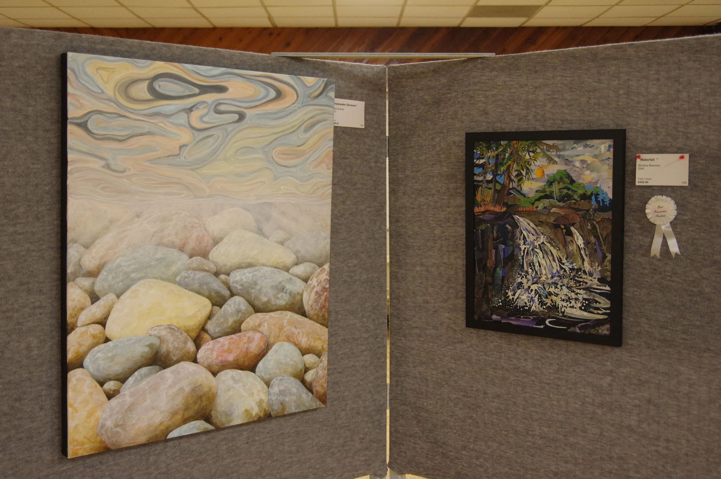 Two framed landscape paintings displayed on gray display boards, one depicting a pile of rocks and swirling clouds, the other showing a waterfall with trees and mountains in the background.