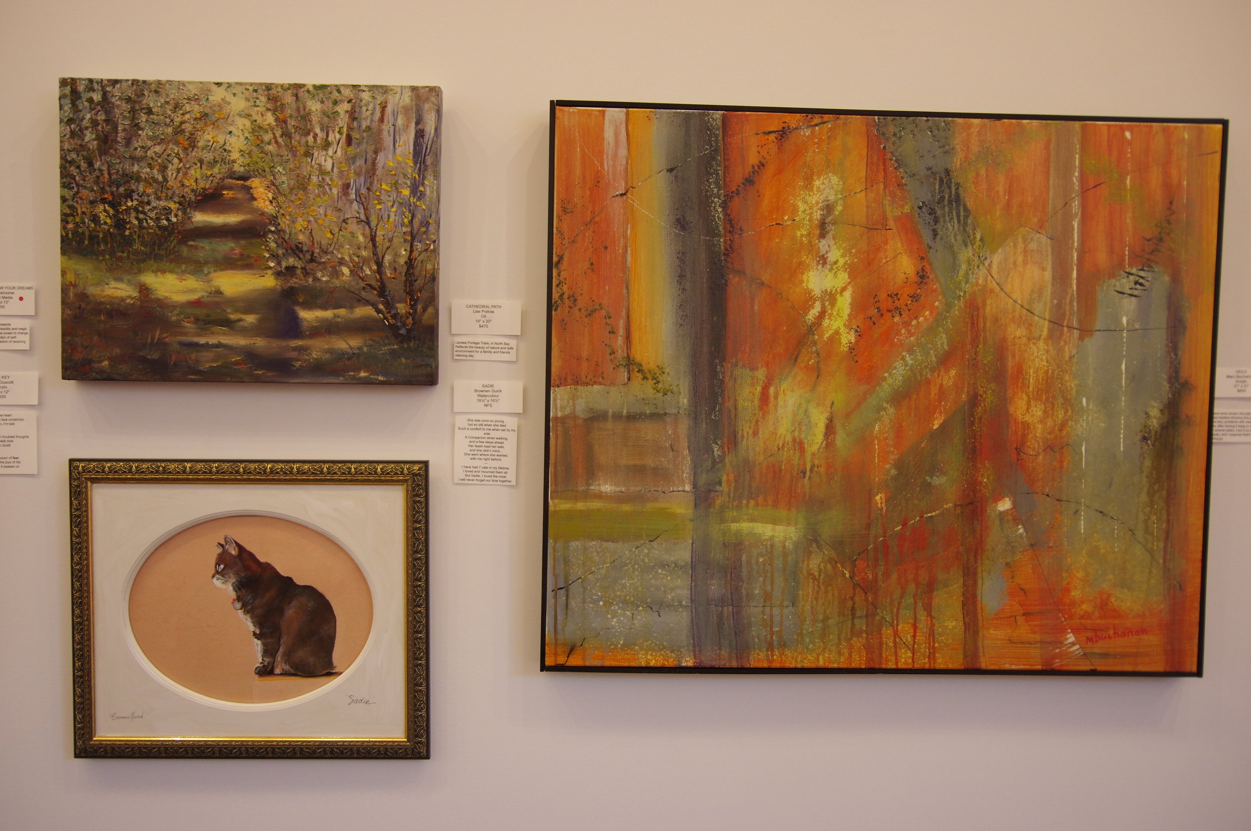 Four paintings on a white gallery wall, including an autumn landscape, an abstract orange and gray artwork, a framed portrait of a brown cat, and a small part of a text panel on the right.