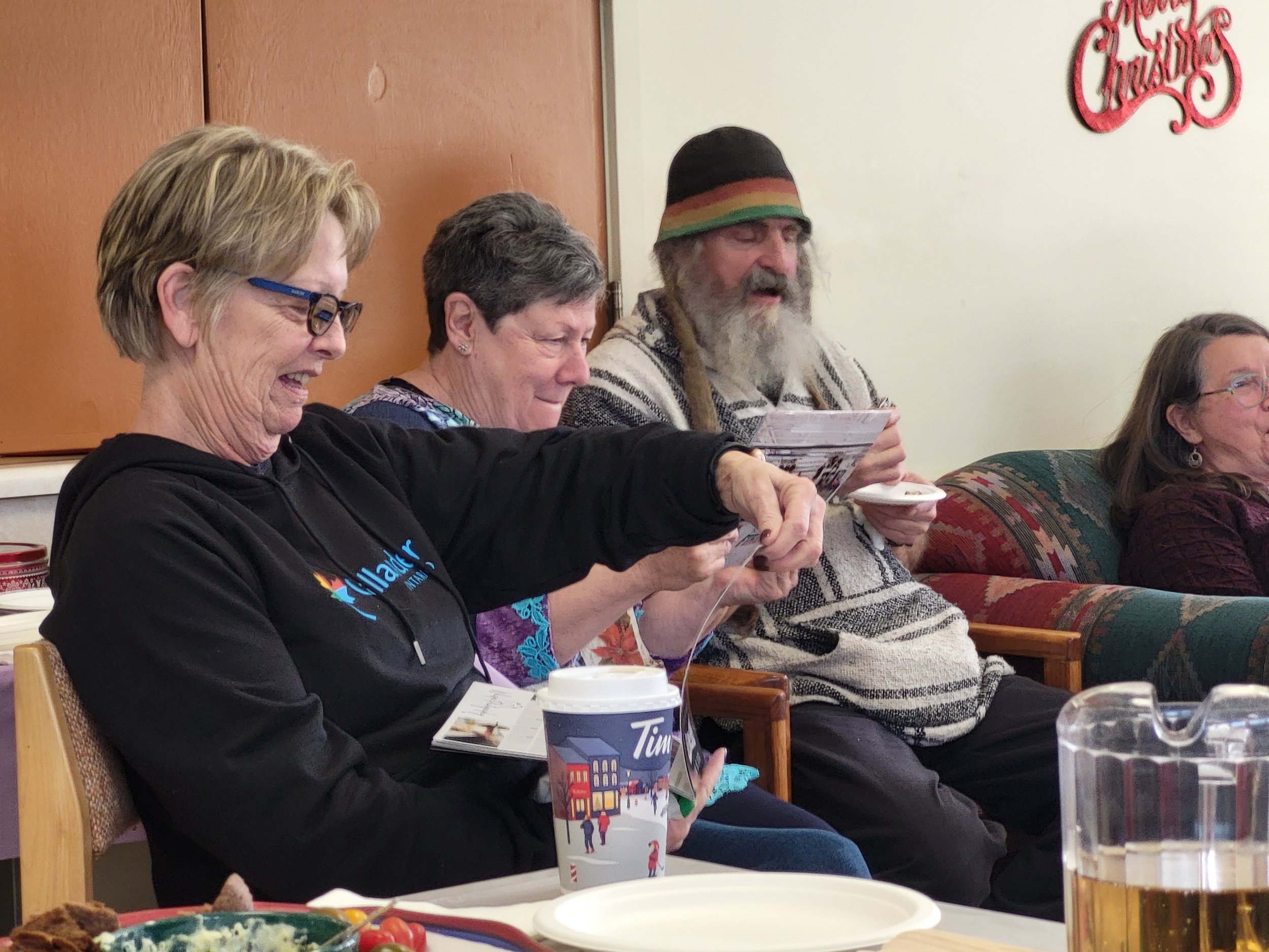 Four people sitting on a couch and chair, smiling and engaging in a social gathering. They are surrounded by food and drinks.