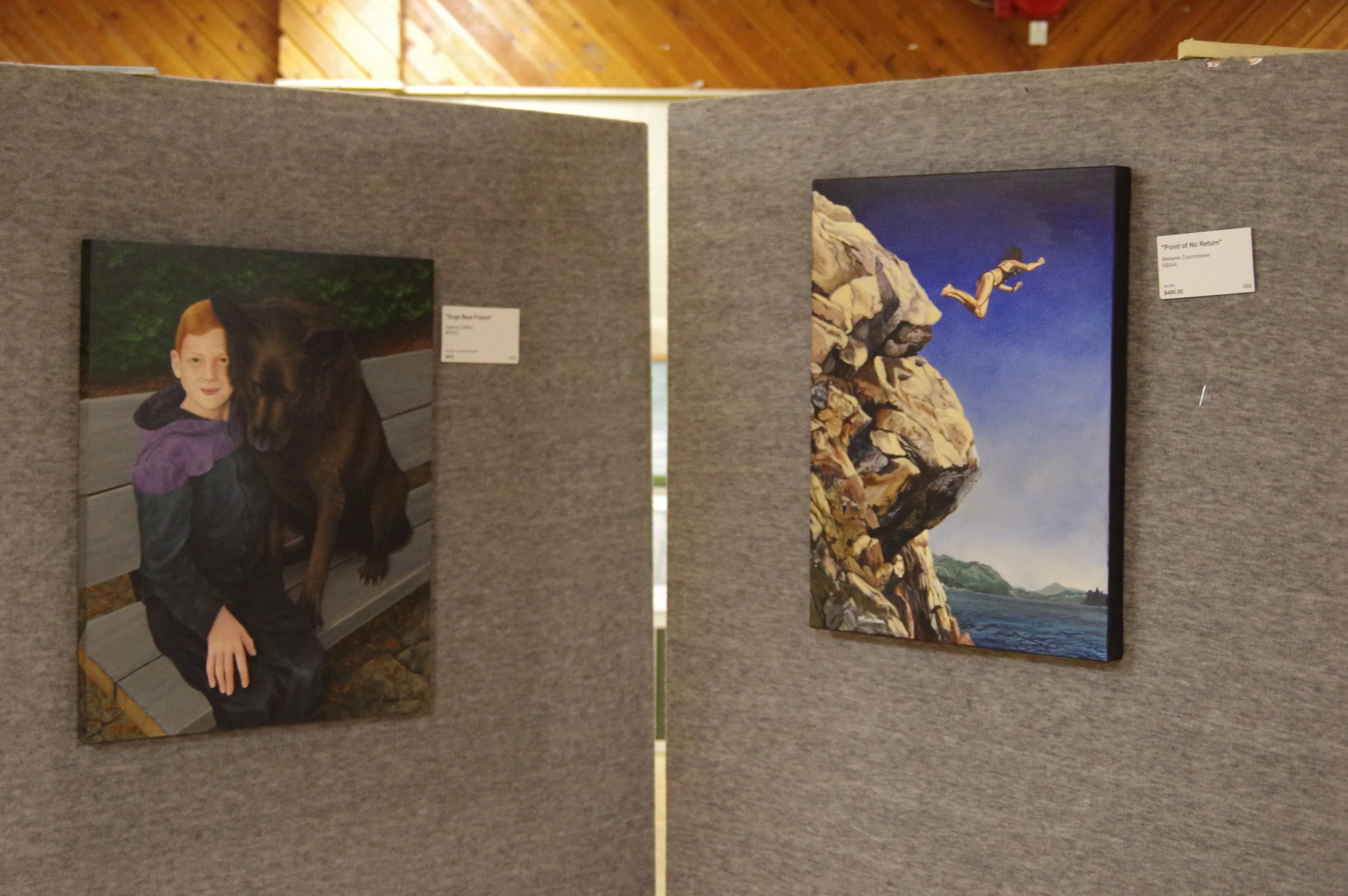 Photograph of a boy with red hair and a black and purple jacket sitting on a wooden bench next to a large black dog, with a rocky nature background, displayed on gray fabric-covered display board. To the right, a painting of a limestone cliff by the ocean with a person diving off the cliff into the water, against a blue sky, listed as "Point of No Return" with a price of $400.00.
