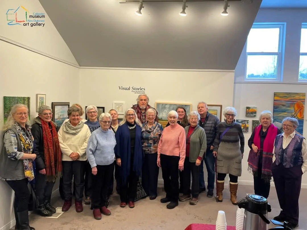 A group of seniors standing together for a photo at an art gallery, with paintings on the walls behind them.