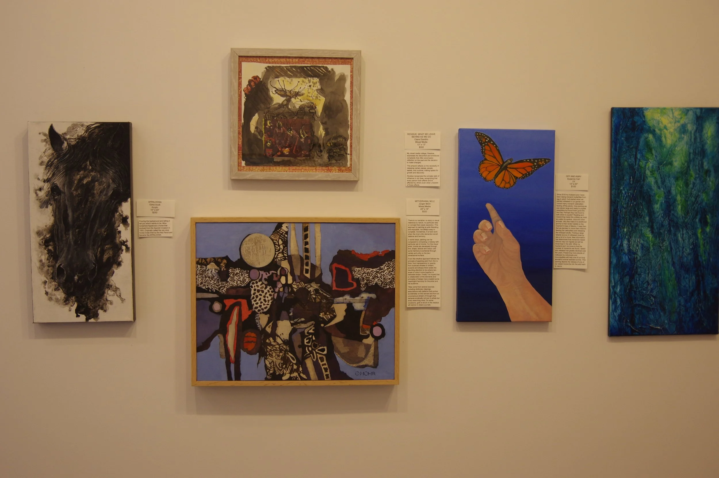Art gallery wall with six paintings displayed, including a black and white horse portrait, abstract landscapes, a hand reaching out to a butterfly, and a textured blue-green artwork.