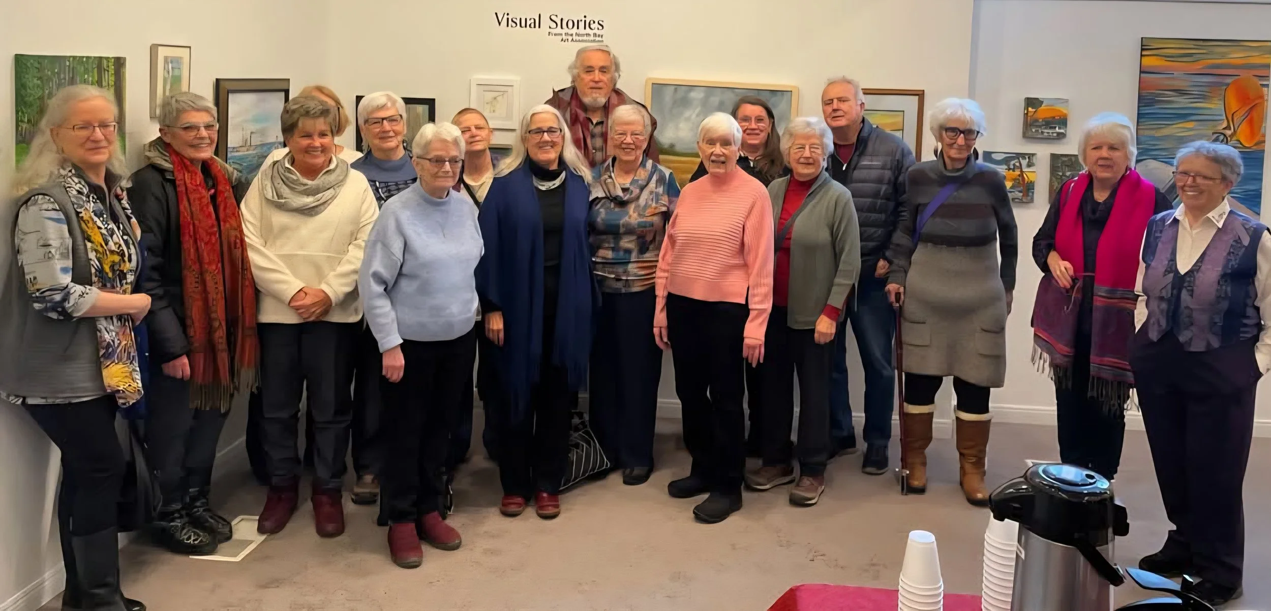 Group of fifteen older adults standing together in an art gallery, smiling, with paintings hanging on the wall behind them.
