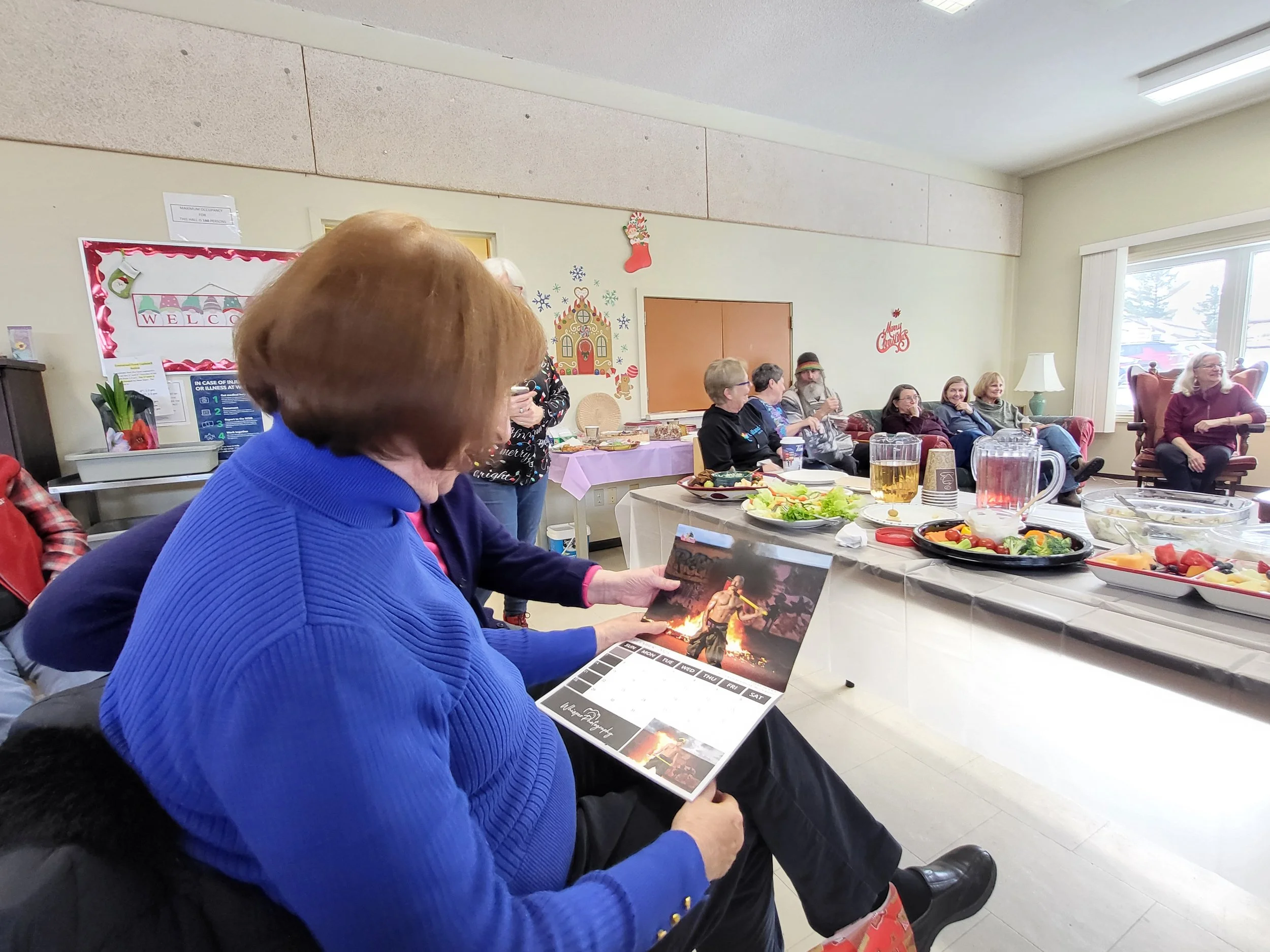A woman in a blue sweater reading a calendar at a holiday gathering with food and drinks on the table and people sitting in chairs in the background.