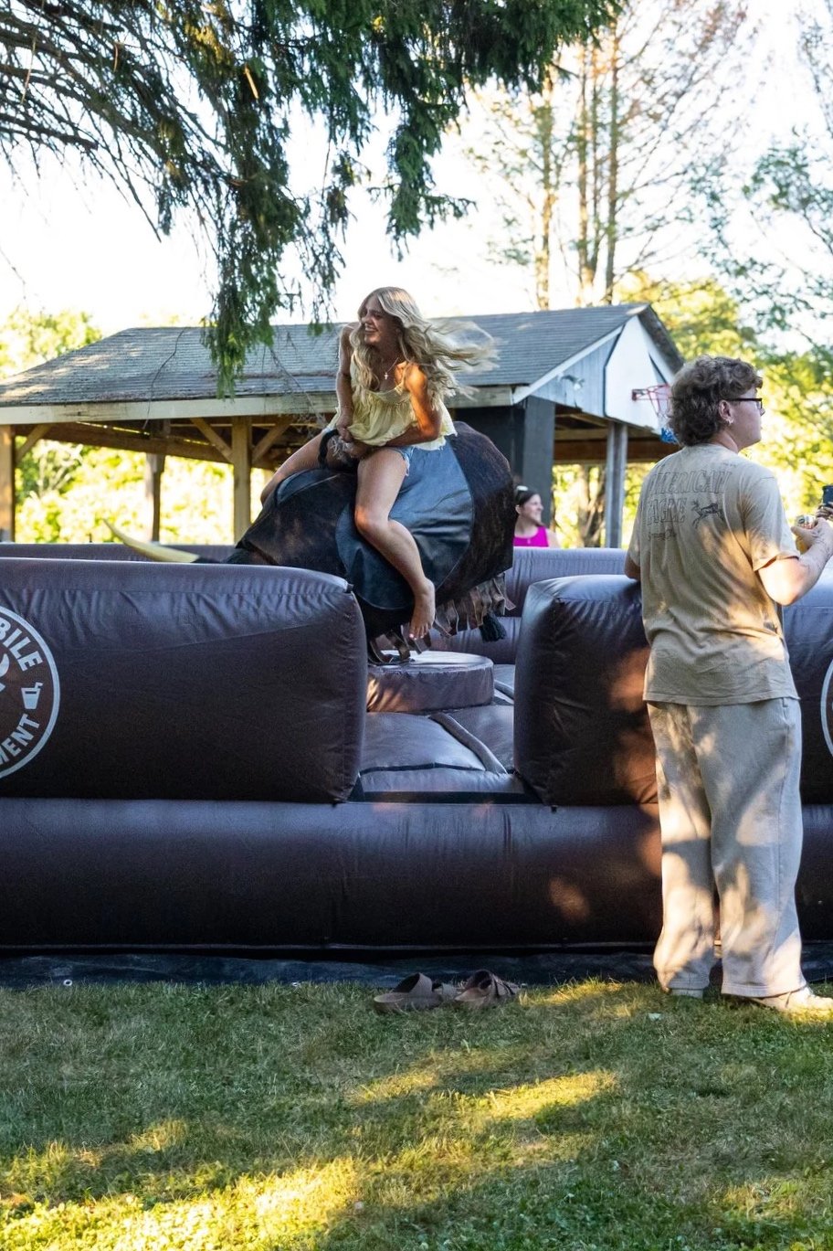 A woman riding a mechanical bull outdoors, with trees and a shed in the background. The woman is smiling and appears to be having fun, and there are people standing nearby.
