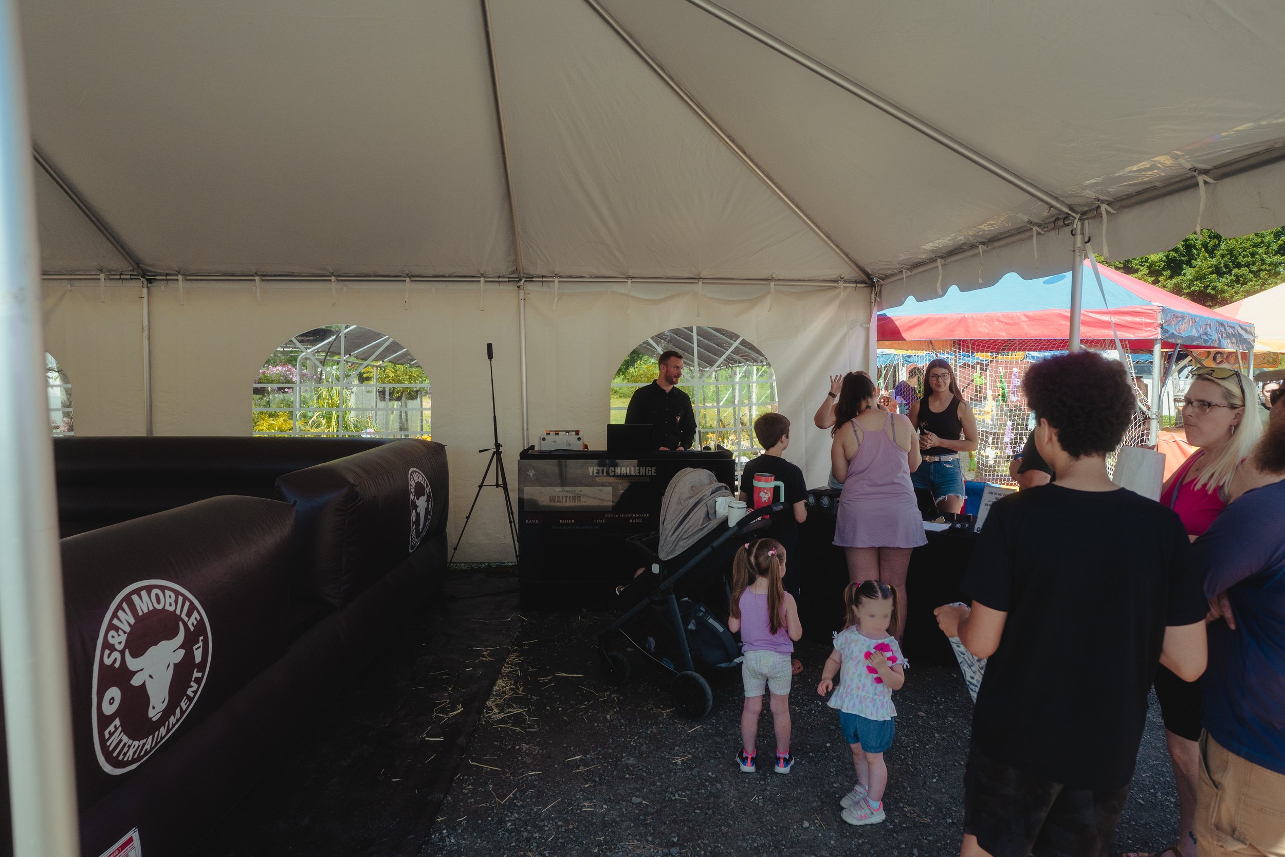 People gathered inside a large white event tent with open sides, with children and adults standing and talking. There are colorful tents and greenery visible outside. The tent has black banners with white logos, and a DJ is playing music with equipment labeled 'YETI CHALLENGE'.