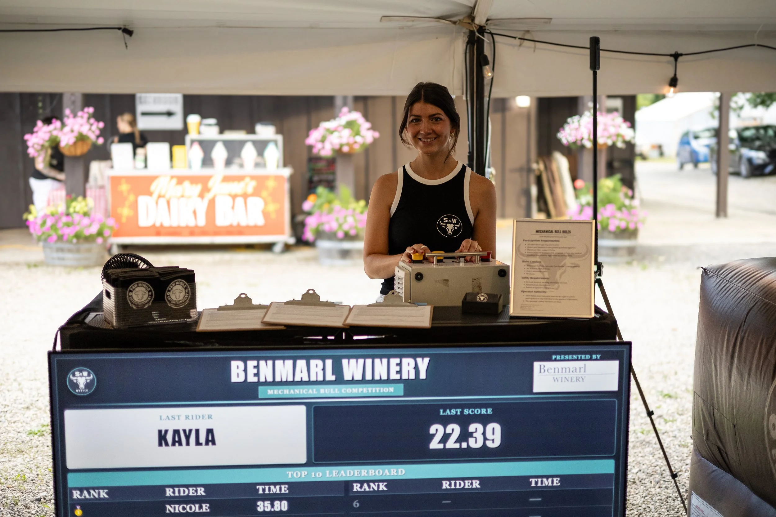 A woman stands at a table with a mechanical bull riding competition scoreboard for Benmarl Winery. The scoreboard and mechanical bull are operated by S&W Mobile Entertainment.
